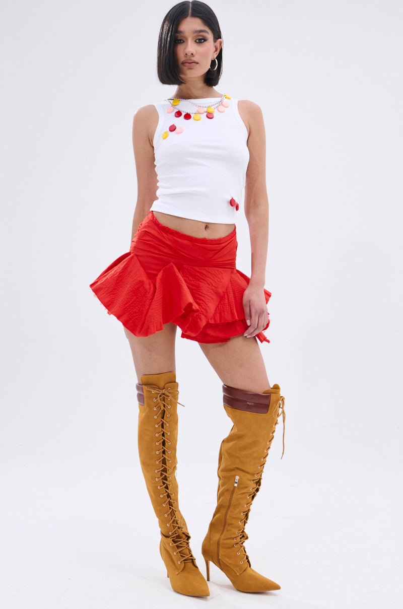 A woman poses against a plain background wearing the GIRL POWER TANK—a white ribbed knit tank top with colorful circular decorations—paired with a red ruffled mini skirt and tall tan lace-up high-heeled boots.