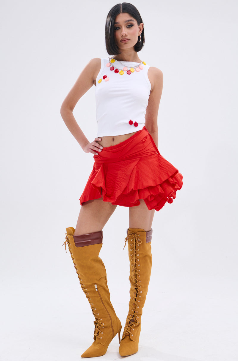 A woman confidently poses against a plain white background wearing the GIRL POWER TANK, featuring ribbed knit fabric and colorful pom-poms, paired with a red ruffled mini skirt and tall mustard-yellow lace-up boots.