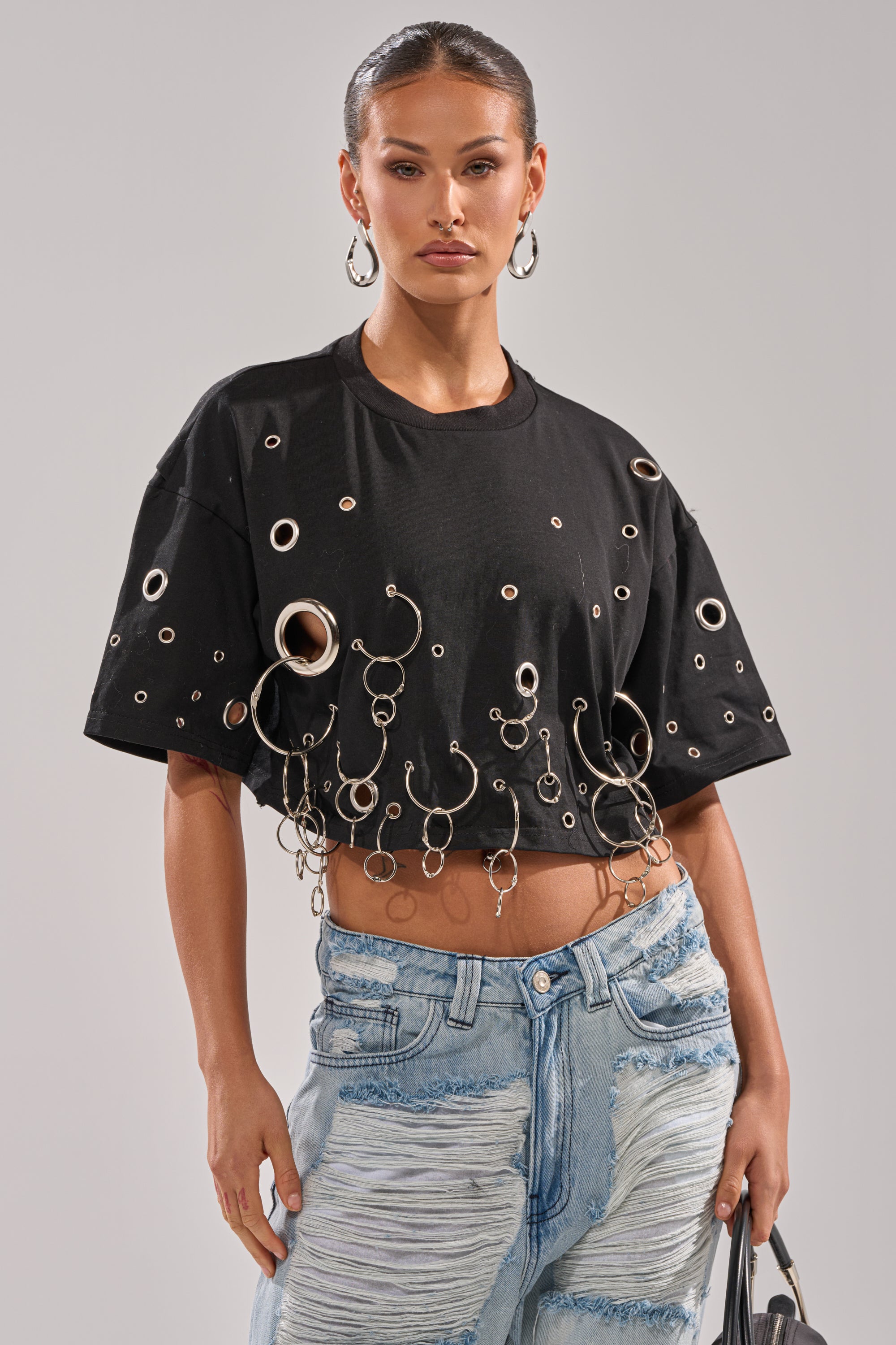 She wears the RING AROUND ME CROPPED TEE with silver grommet and ring details, paired with light blue distressed jeans, hoop earrings, slicked-back hair, and carries a black handbag by her side. 0