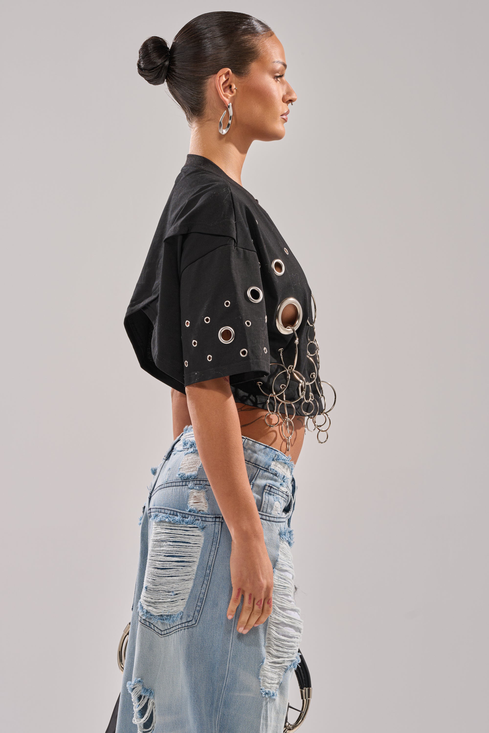 A woman in profile wears the RING AROUND ME CROPPED TEE, a black top with silver grommet and chain accents, styled with distressed light blue jeans, a sleek bun, and large hoop earrings. 2