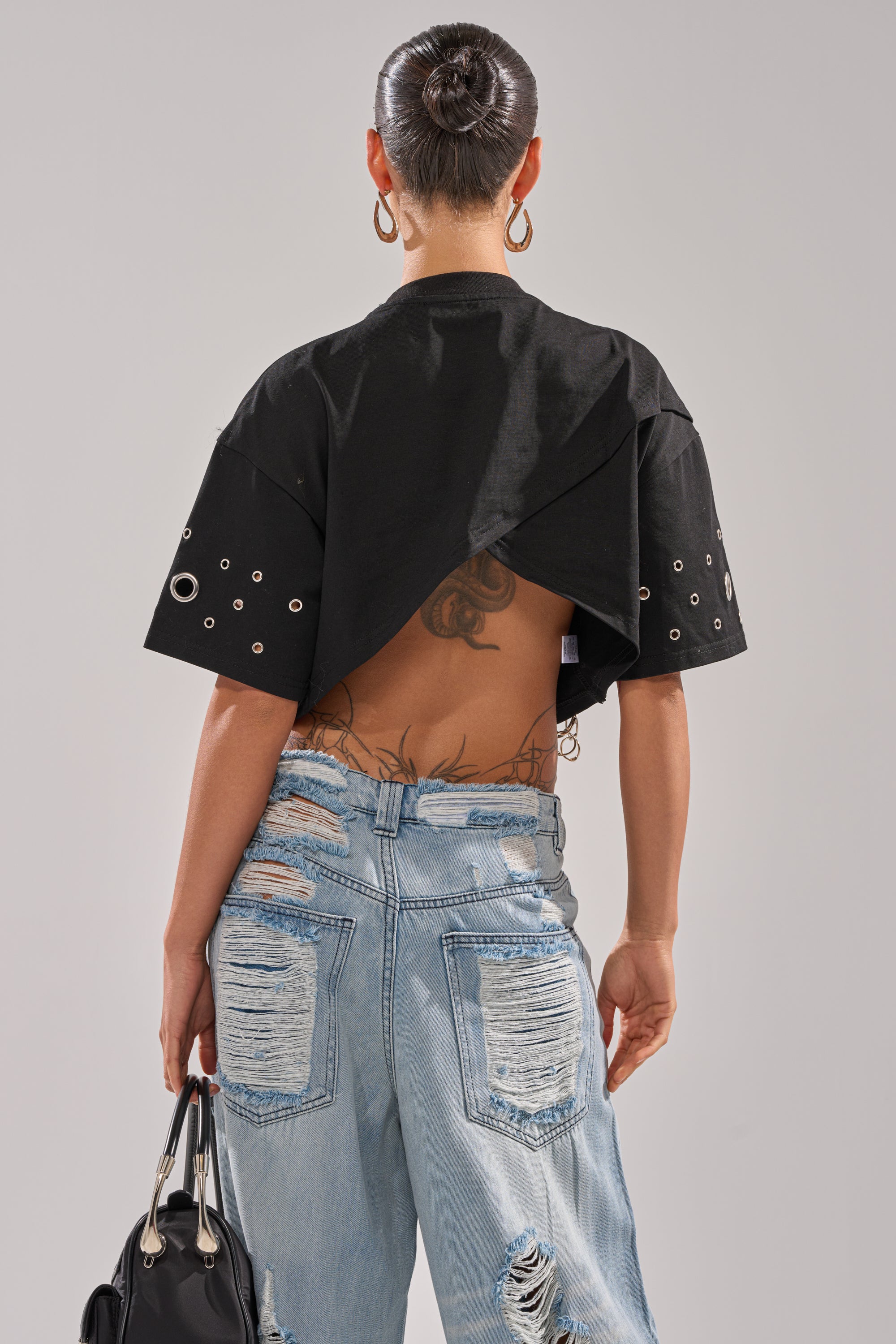 Seen from behind, a person with a sleek bun and hoop earrings wears the RING AROUND ME CROPPED TEE—black with silver grommet details and an open back showing tattoos—paired with light blue ripped jeans and a black handbag. 3