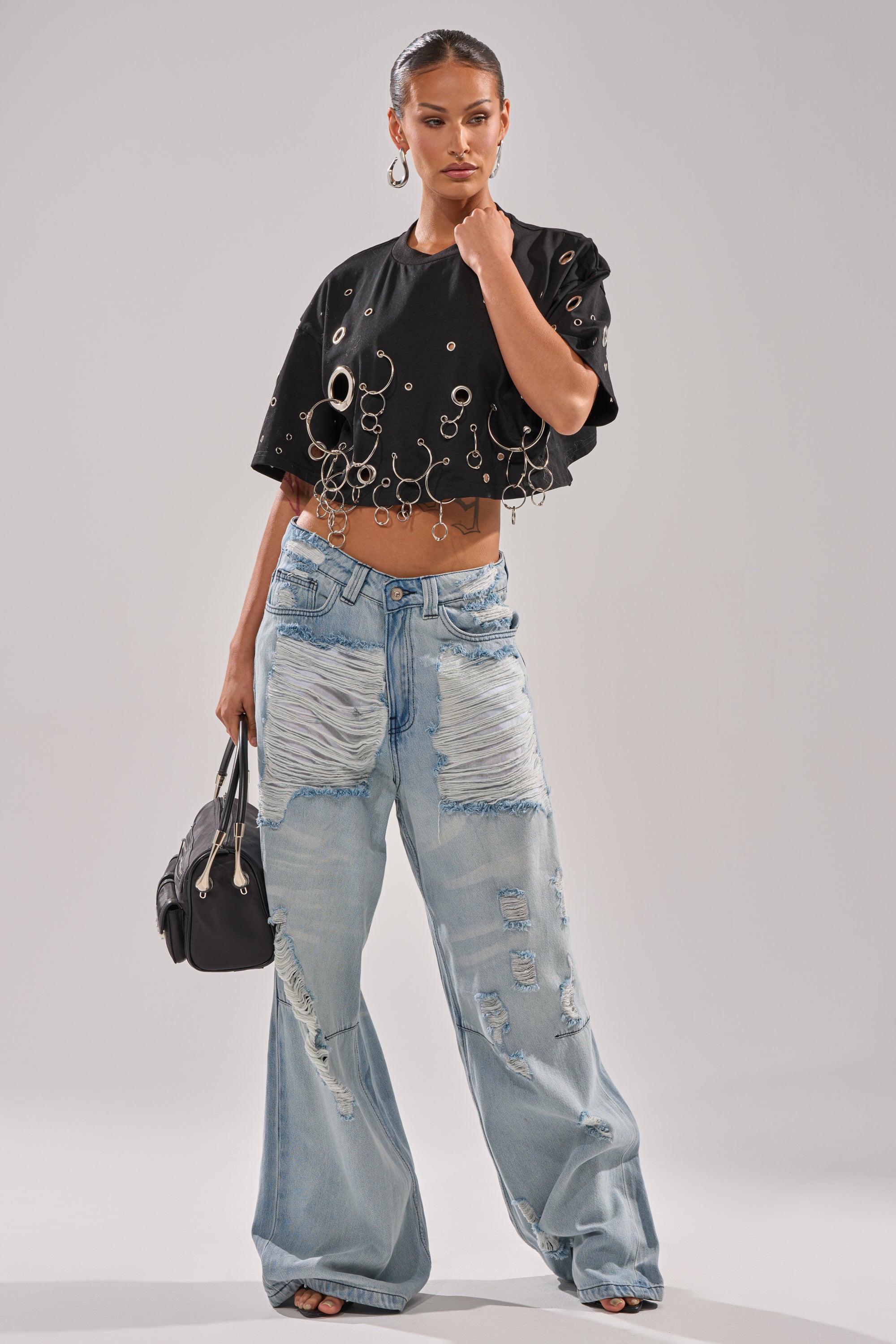 A woman models the RING AROUND ME CROPPED TEE, a black top with silver grommet details, paired with oversized distressed light blue jeans. She accessorizes with a black handbag, black heels, and has her hair sleekly pulled back. 4
