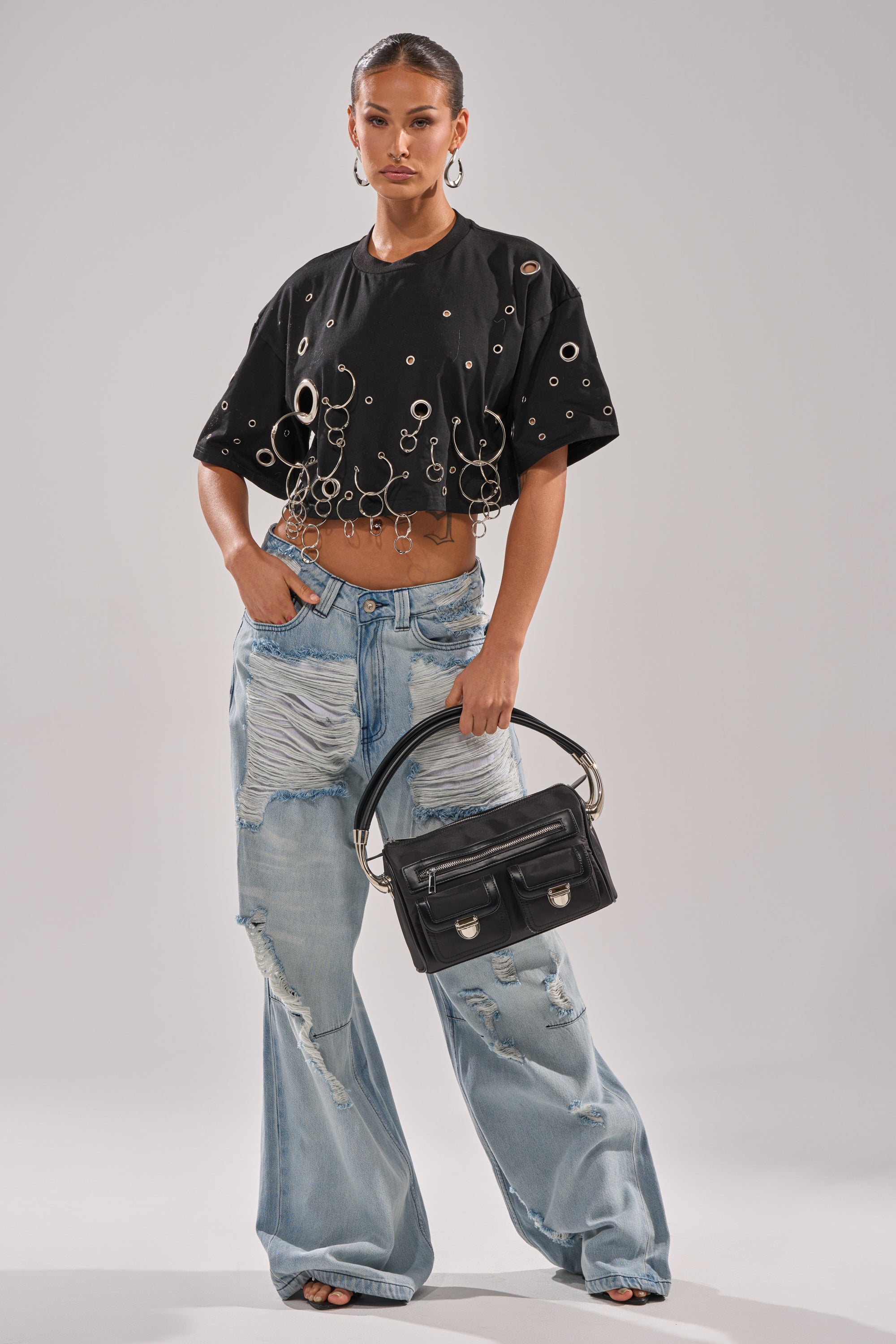 A woman with slicked-back hair and a neutral look stands confidently in the RING AROUND ME CROPPED TEE, featuring silver grommet details and chains, paired with distressed baggy jeans and holding a black handbag by the handle. 5