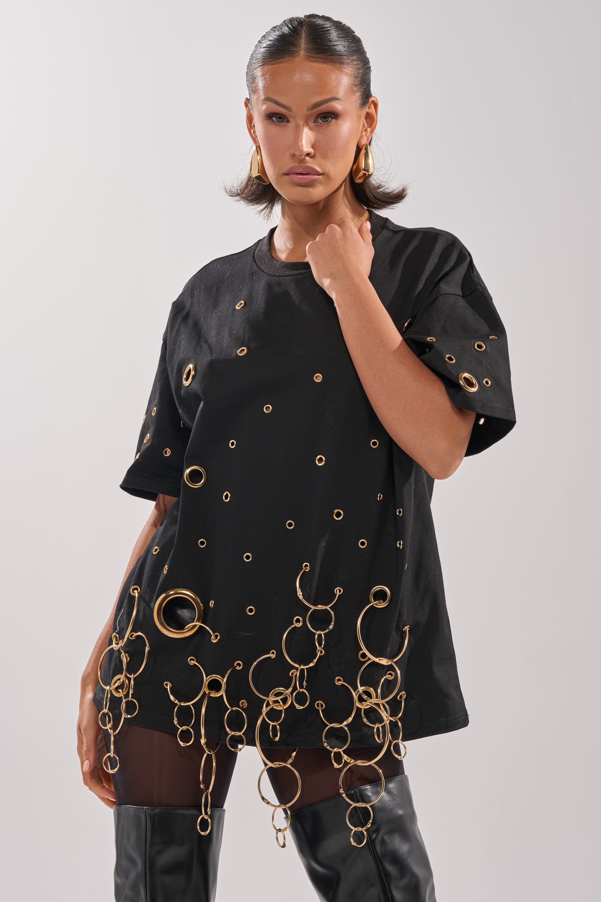 She wears the RING AROUND ME OVERSIZED TEE, featuring metallic grommets and gold rings at the hem, styled with large gold hoop earrings and thigh-high black boots for bold streetwear flair.