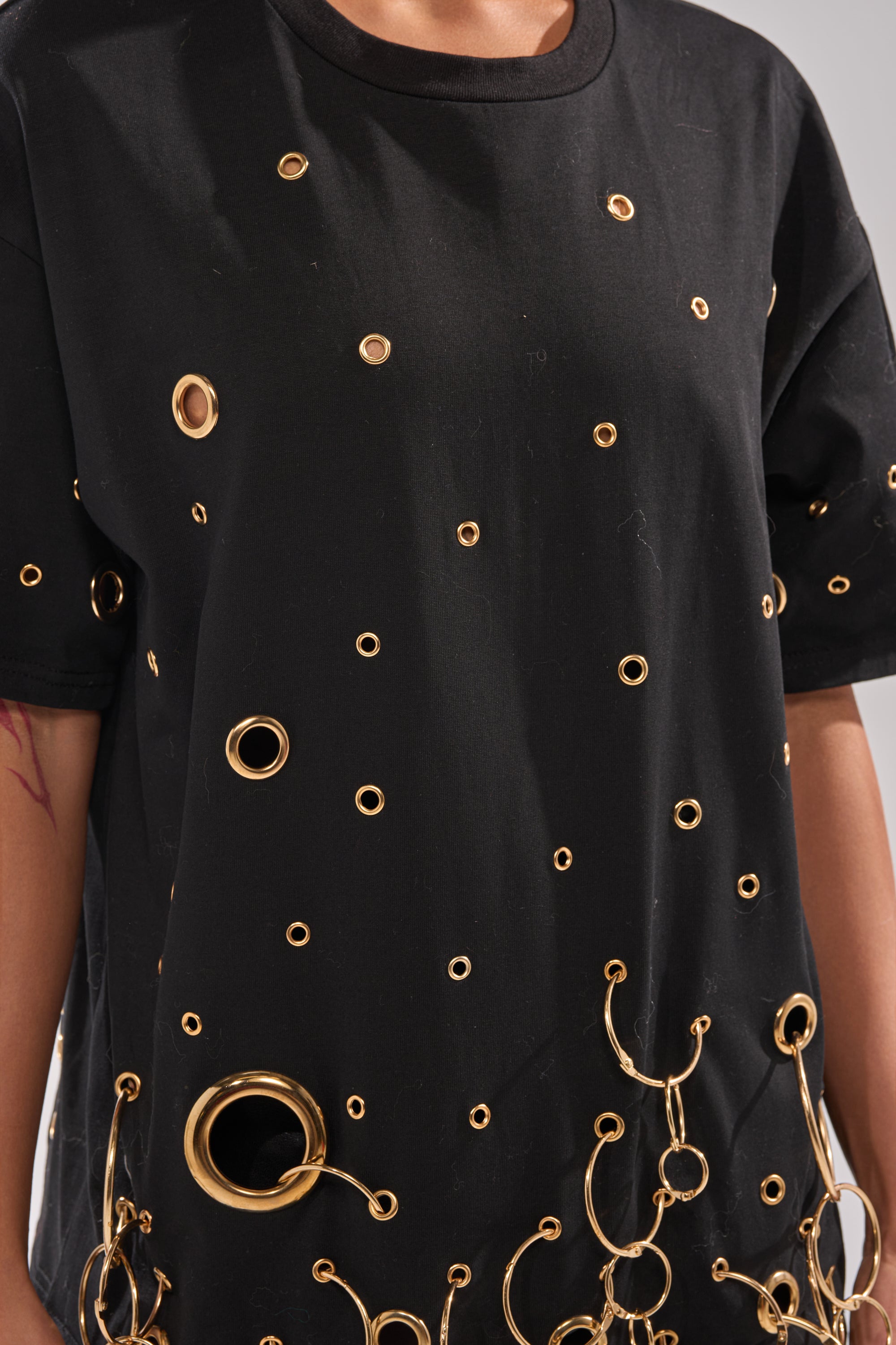 A person wears the RING AROUND ME OVERSIZED TEE, a black streetwear-inspired top featuring an array of metallic grommets and rings in a scattered design. Only the upper body is shown.