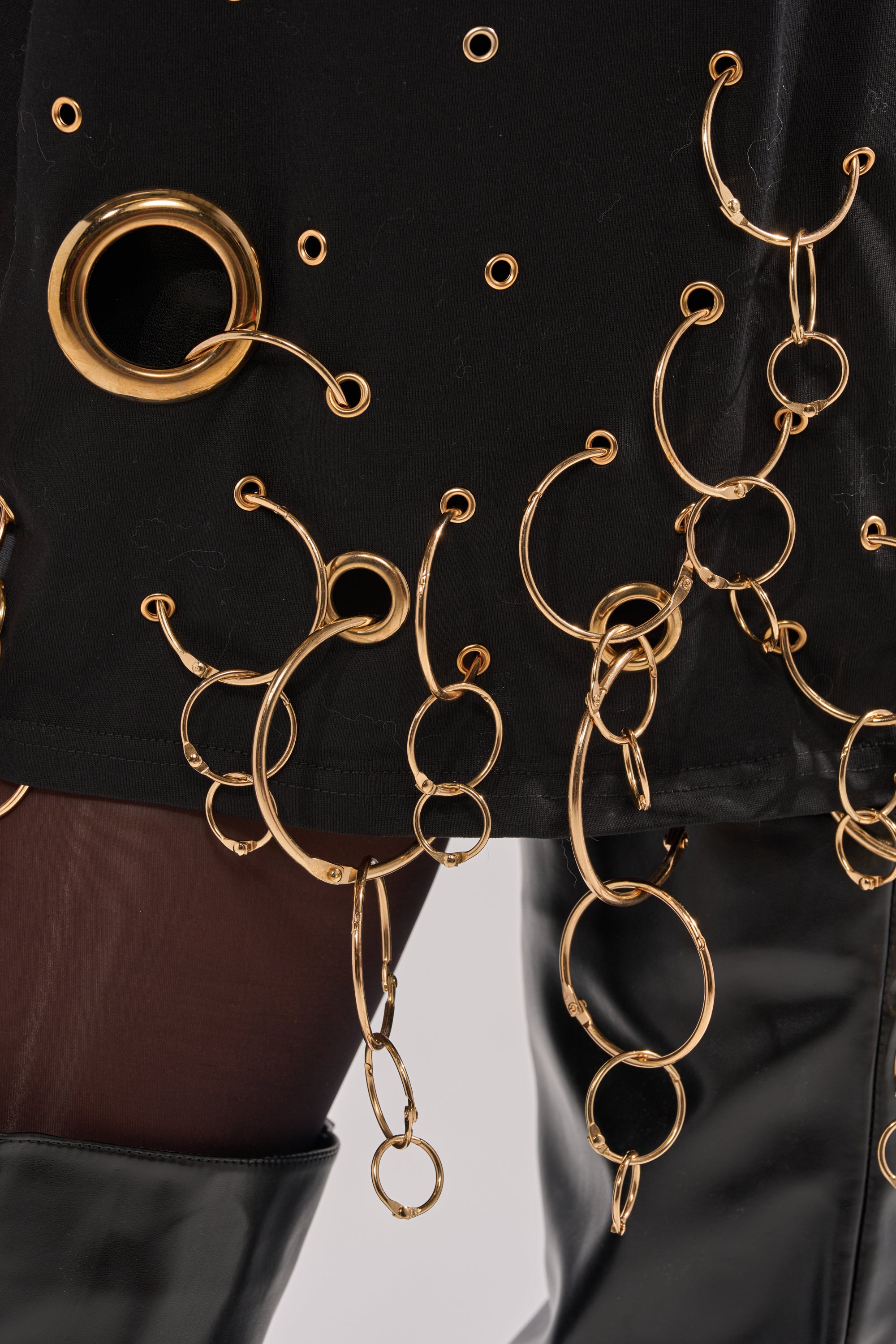 Close-up of the RING AROUND ME OVERSIZED TEE, a streetwear-inspired black top decorated with metallic grommets and gold rings, styled with sheer brown tights and shiny black boots.