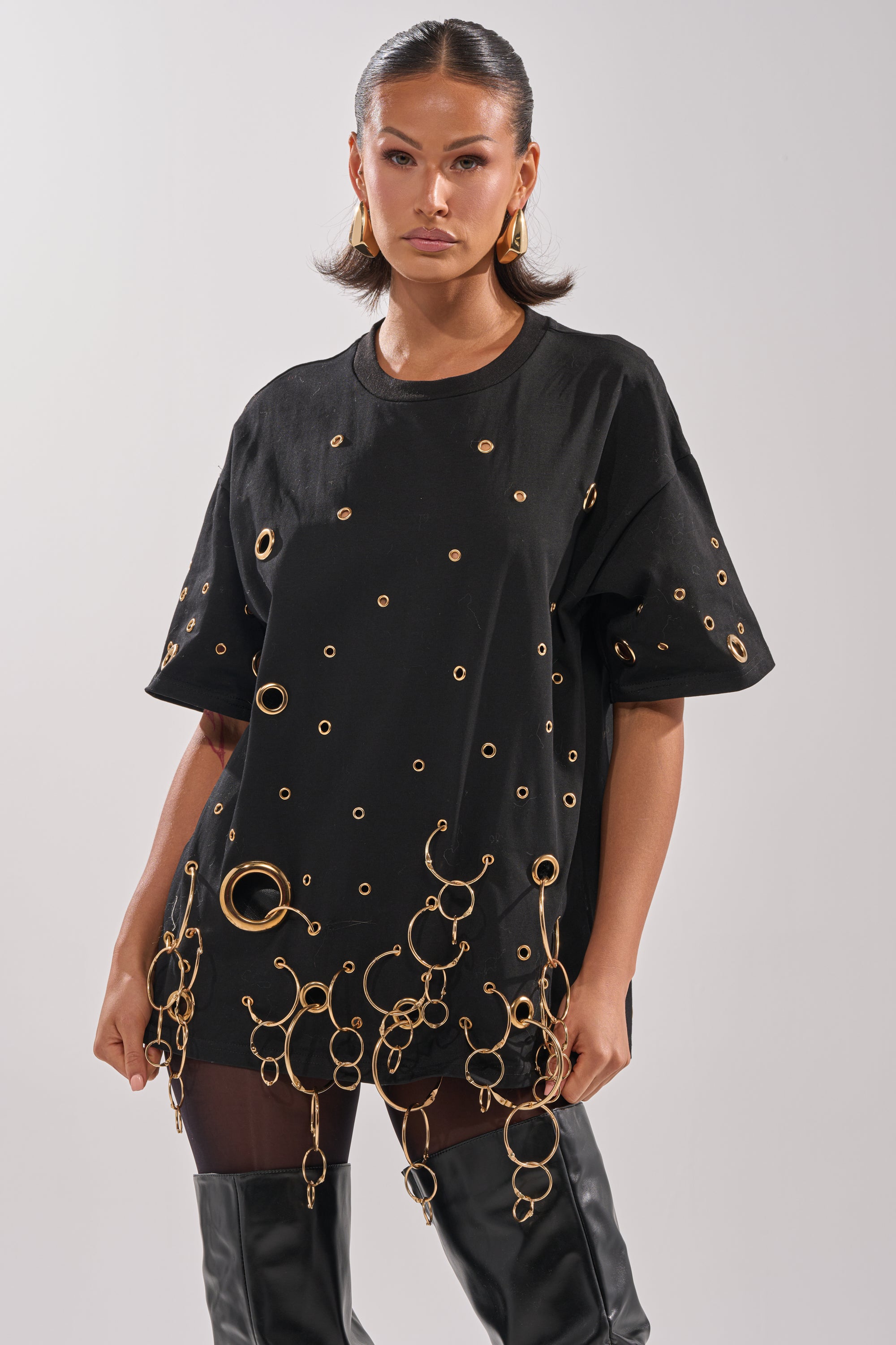 A woman in the RING AROUND ME OVERSIZED TEE, detailed with metallic grommets and gold rings, pairs it with thigh-high black boots against a plain light background for a striking streetwear vibe.