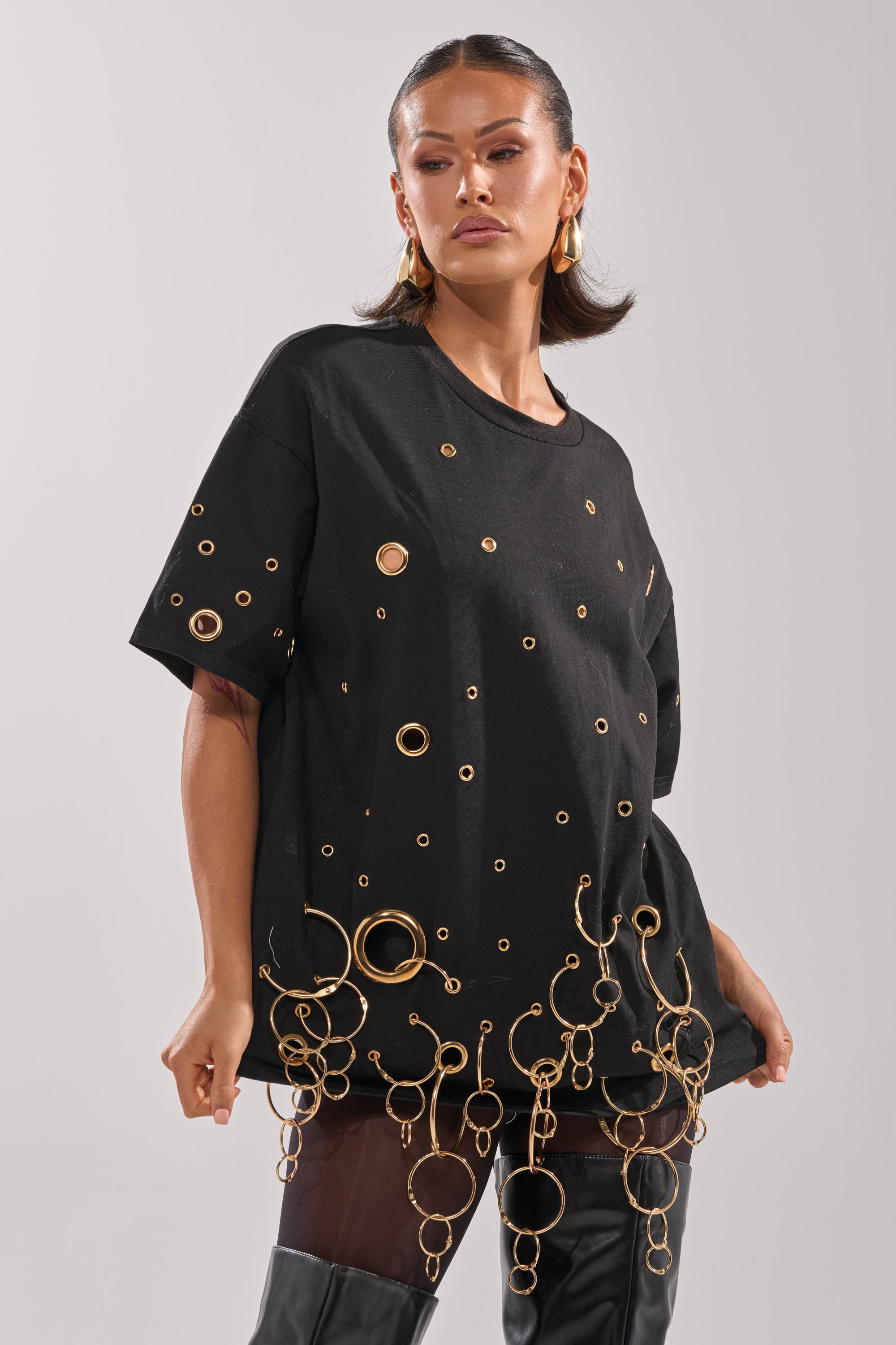 Wearing the RING AROUND ME OVERSIZED TEE with metallic grommets and gold rings at the hem, she pairs it with black thigh-high boots, posing confidently against a plain background for a bold streetwear look.