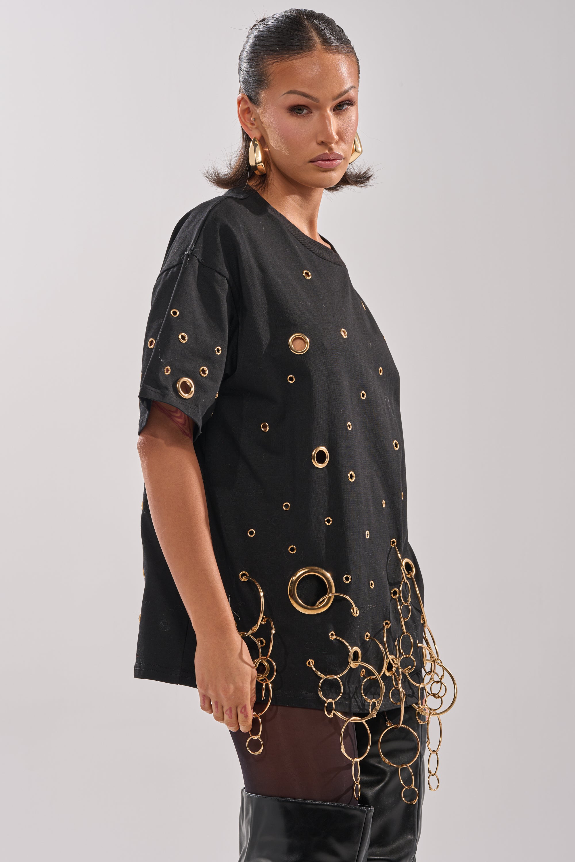 She wears the RING AROUND ME OVERSIZED TEE, featuring metallic grommets and bold gold rings, styled with black thigh-high boots for an edgy, statement streetwear look.
