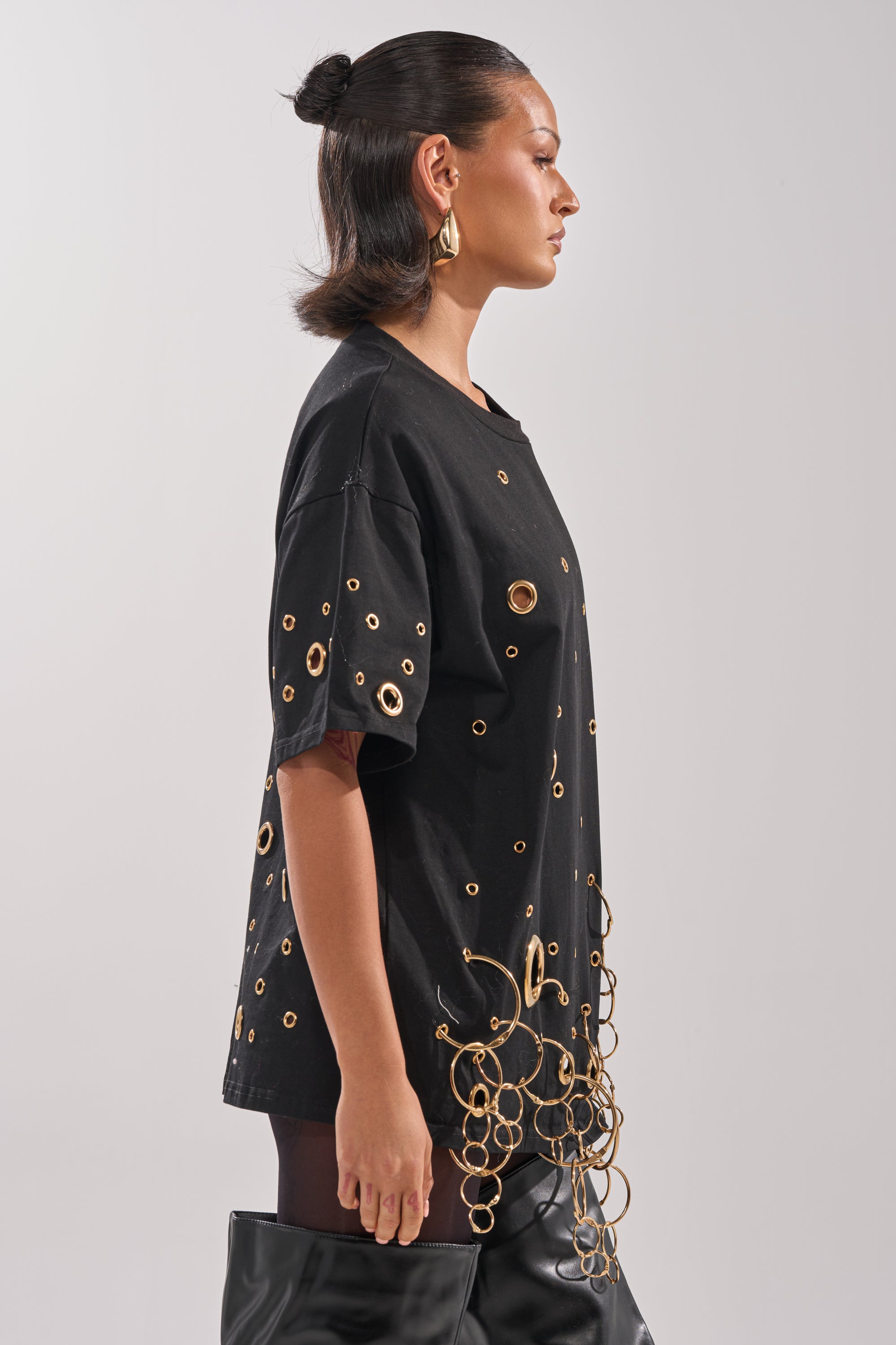 A woman in profile wears the RING AROUND ME OVERSIZED TEE with metallic grommets, paired with black leather boots. Her pulled-back, shoulder-length hair is styled with large gold hoop earrings. The background is plain.