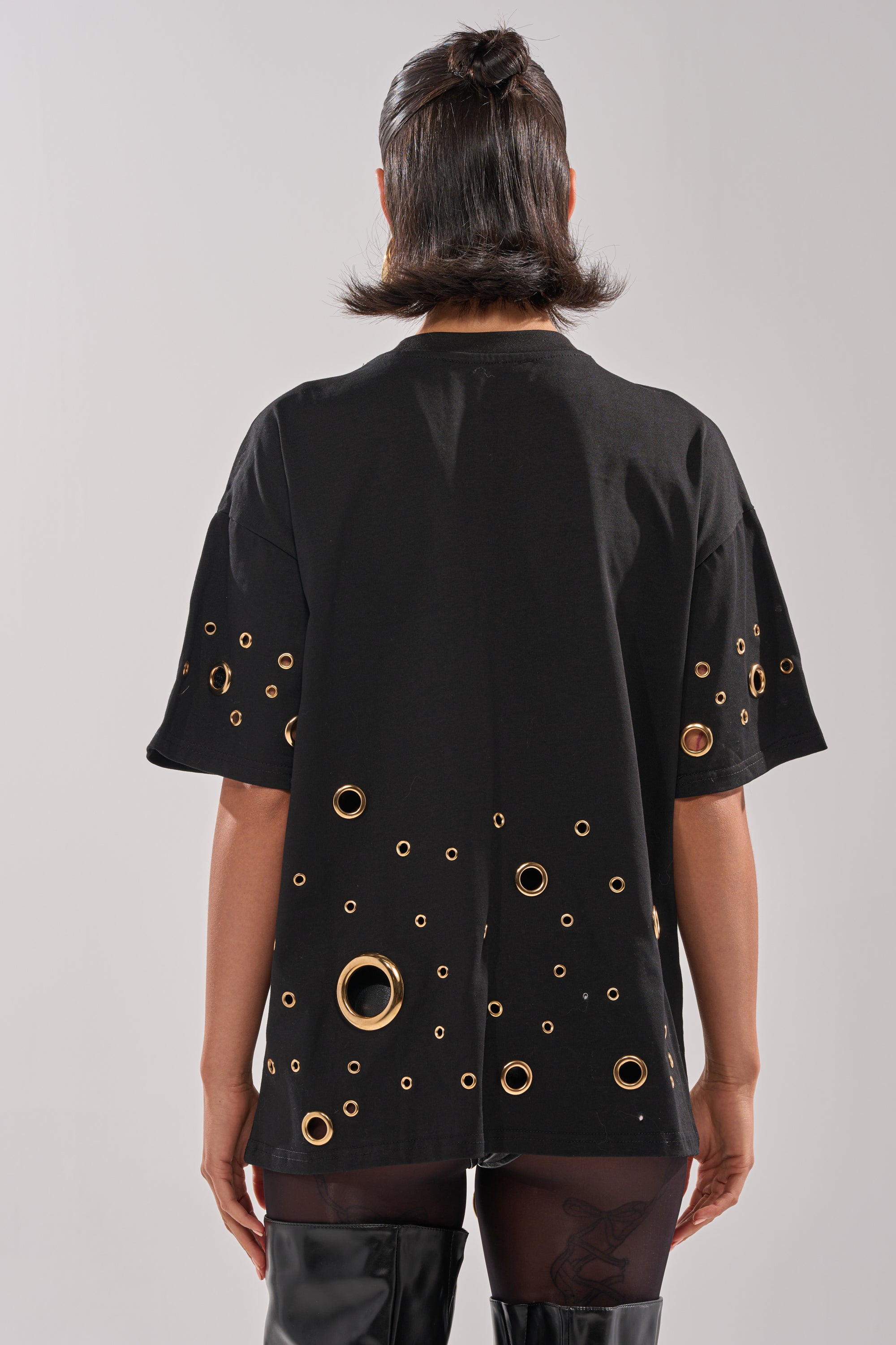 A person faces away, wearing the RING AROUND ME OVERSIZED TEE with metallic grommets on the back and sleeves. They style it with sheer tights and black over-the-knee boots for a bold streetwear look.