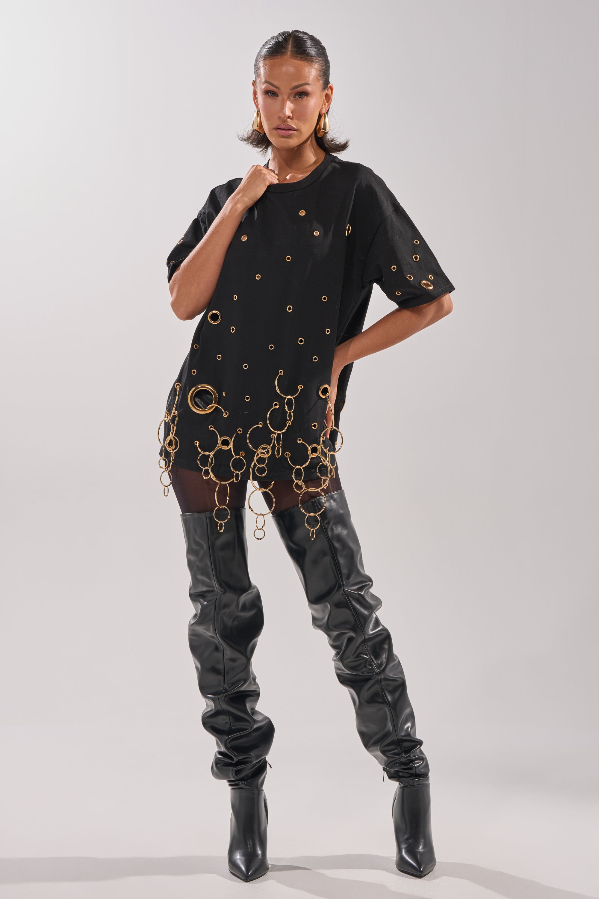 A woman models the RING AROUND ME OVERSIZED TEE styled with gold rings, chains, and thigh-high black leather boots against a light gray background, creating a bold and edgy streetwear look.