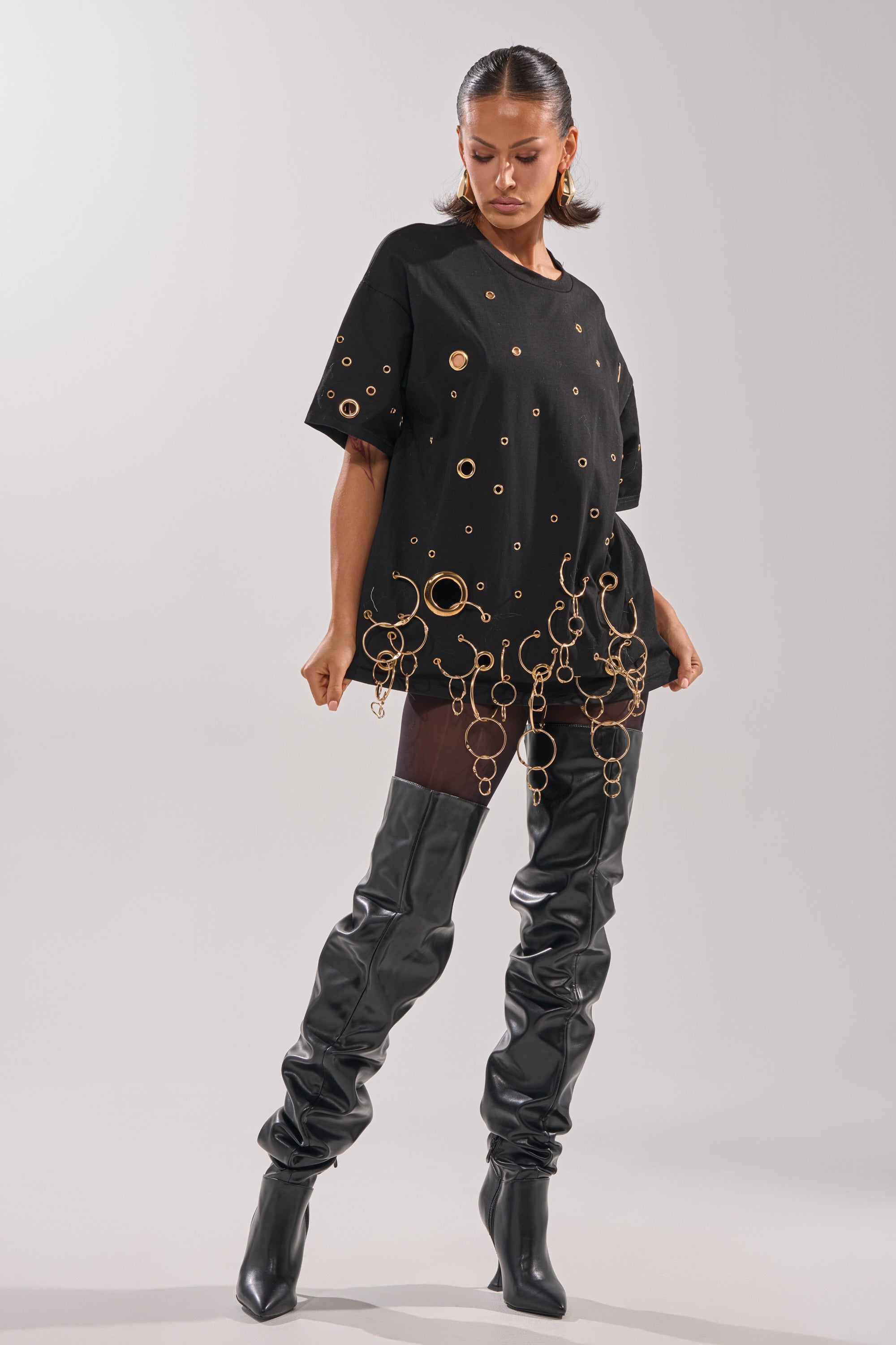 A woman models the RING AROUND ME OVERSIZED TEE, featuring metallic grommets and gold ring details, styled with slouchy black leather thigh-high boots against a plain backdrop for a bold streetwear statement.