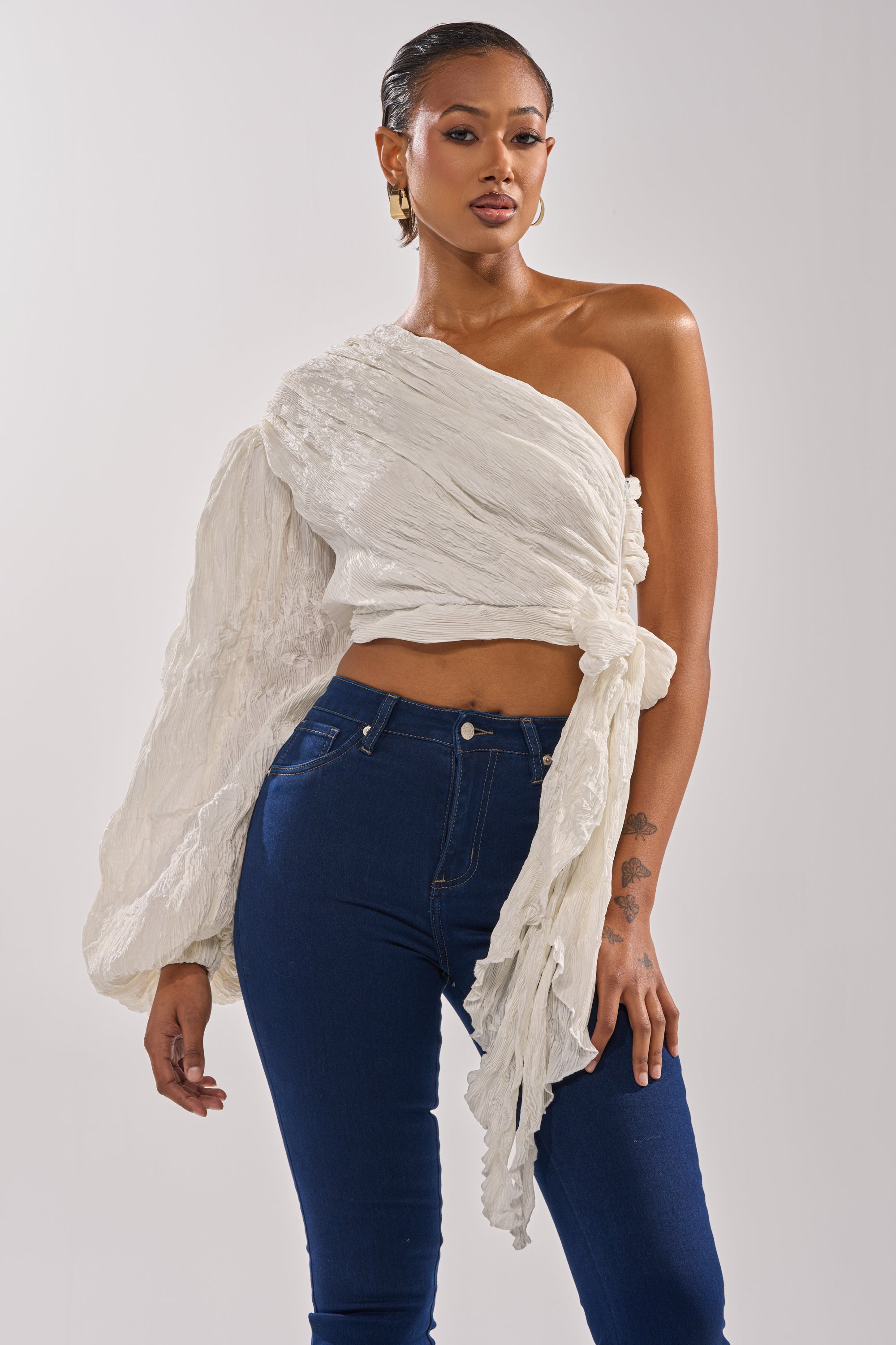 A woman poses confidently in the GROOVY BABY SIDE TIE BLOUSE, featuring textured fabric and a one-shoulder design, paired with dark blue high-waisted jeans. She completes the look with gold hoop earrings, slicked-back hair, and a visible wrist tattoo. 0