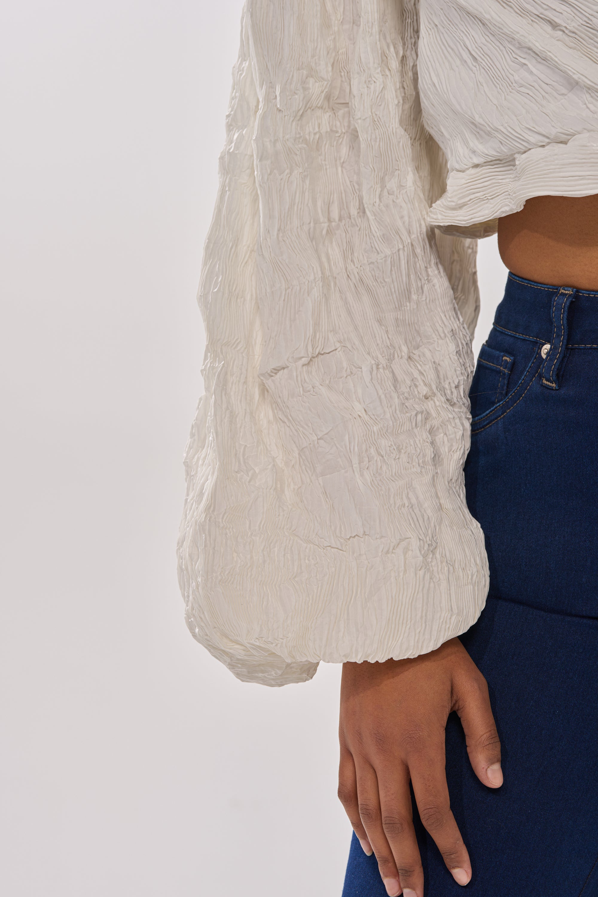 Wearing the GROOVY BABY SIDE TIE BLOUSE, a textured white blouse with billowy sleeves, paired with dark blue high-waisted jeans. Shown from the waist down with one arm relaxed at their side. 9
