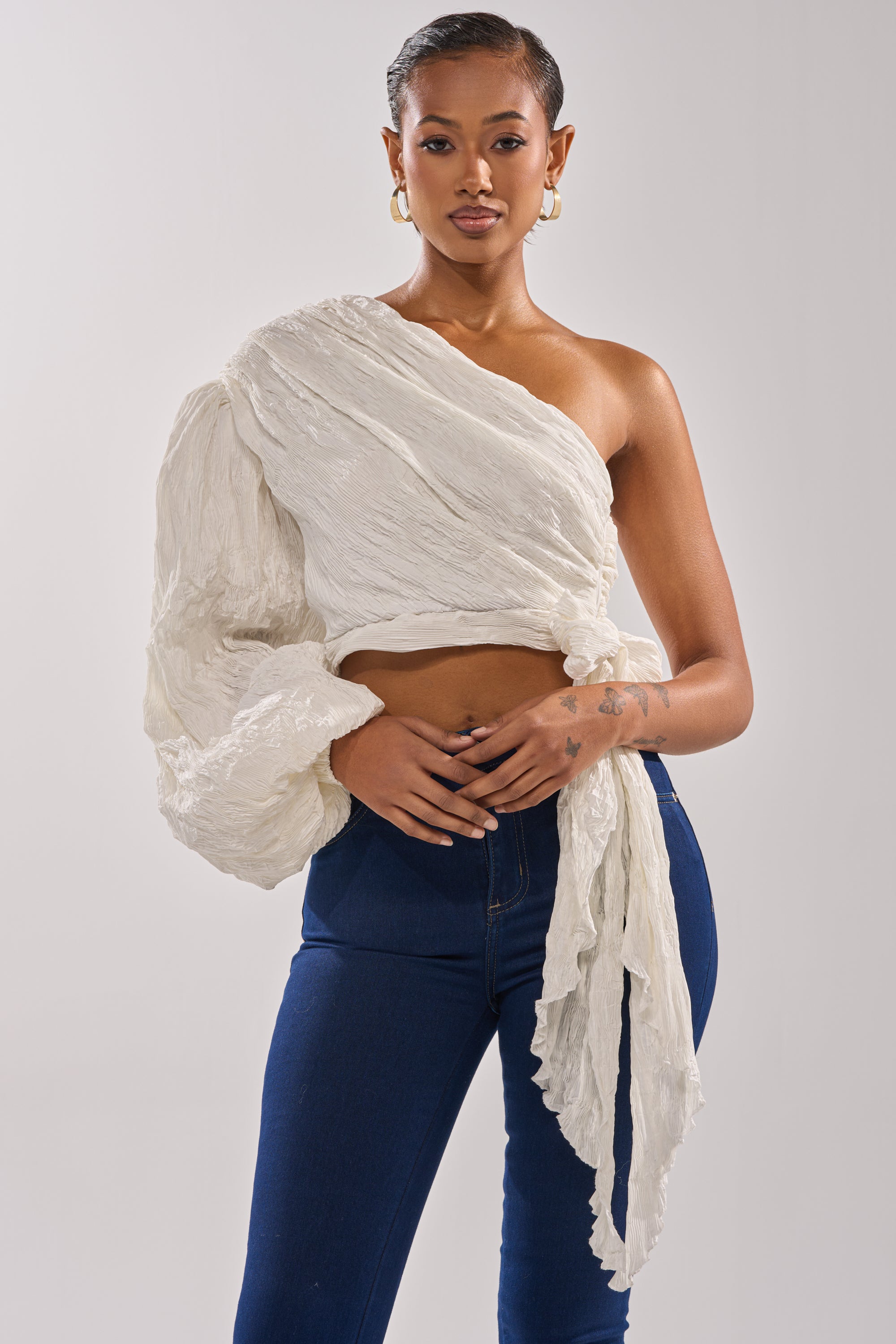 A woman poses confidently in the GROOVY BABY SIDE TIE BLOUSE, a textured off-white one-shoulder top with voluminous sleeves and side tie, paired with dark blue high-waisted jeans against a plain light background. 1