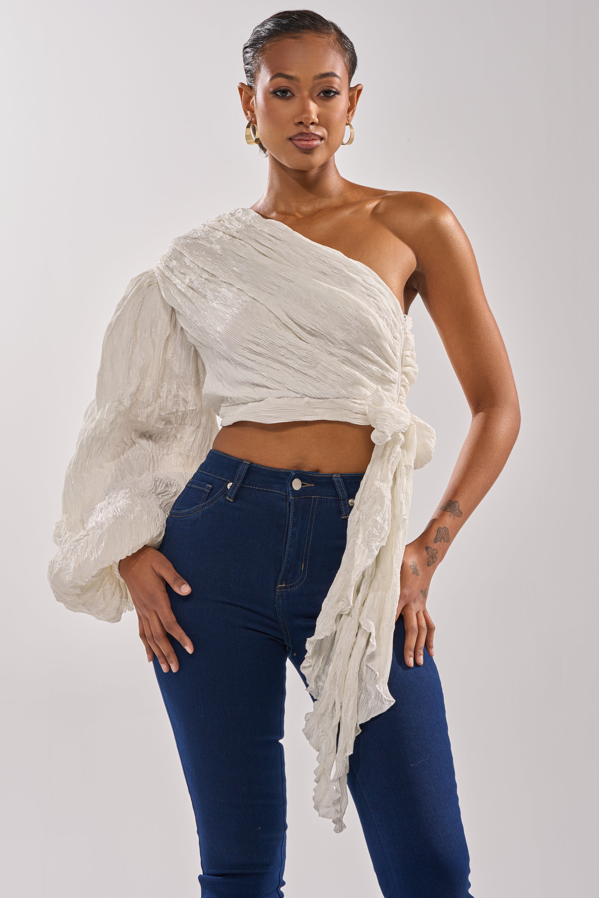 A woman poses confidently in the GROOVY BABY SIDE TIE BLOUSE, a textured off-white one-shoulder top with a dramatic sleeve and side tie at the waist, paired with dark blue high-waisted jeans against a plain background. 2