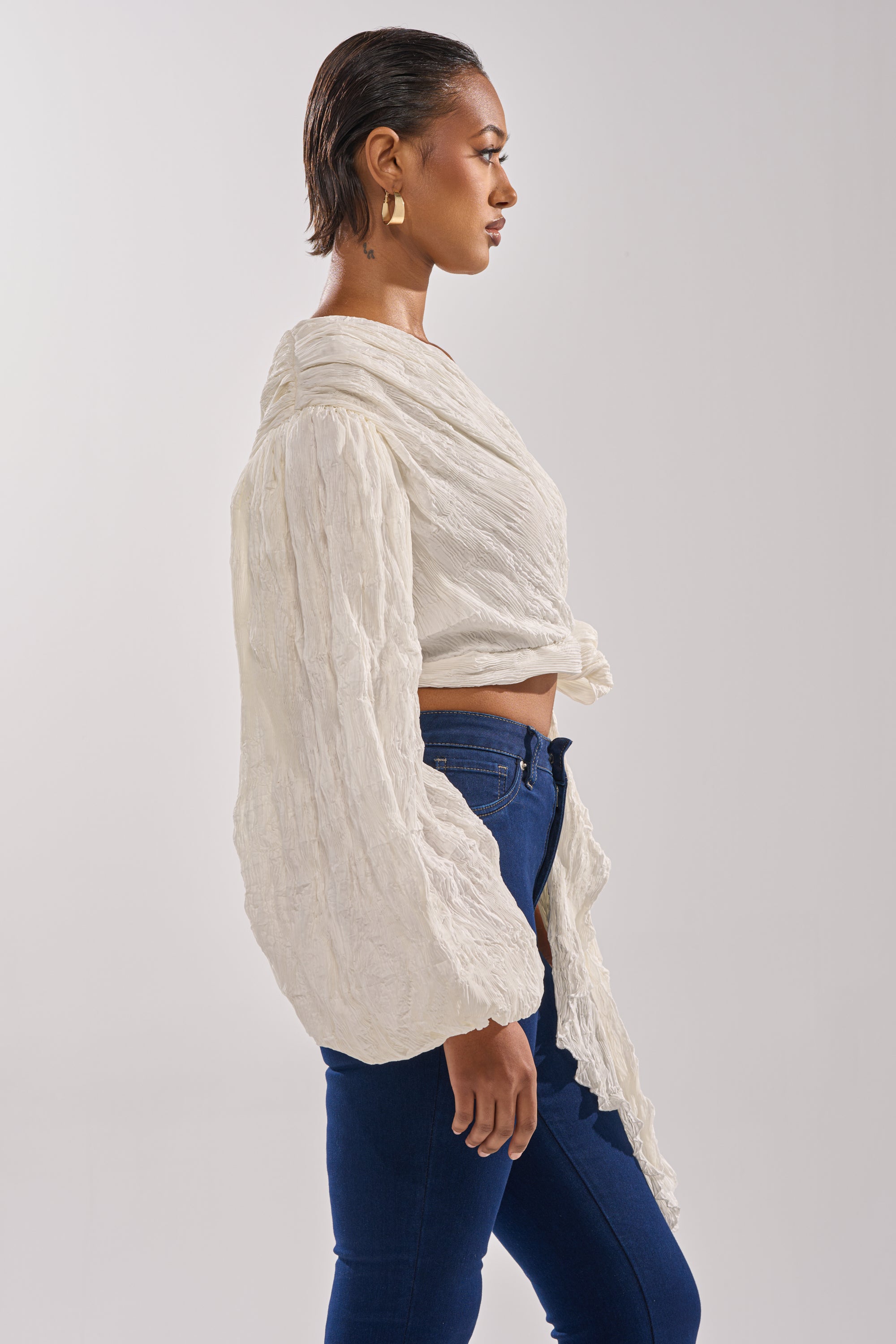 A woman with short hair stands in profile, wearing the GROOVY BABY SIDE TIE BLOUSE—a textured white top with voluminous sleeves and cropped fit—paired with blue jeans and gold hoops against a plain light background. 3