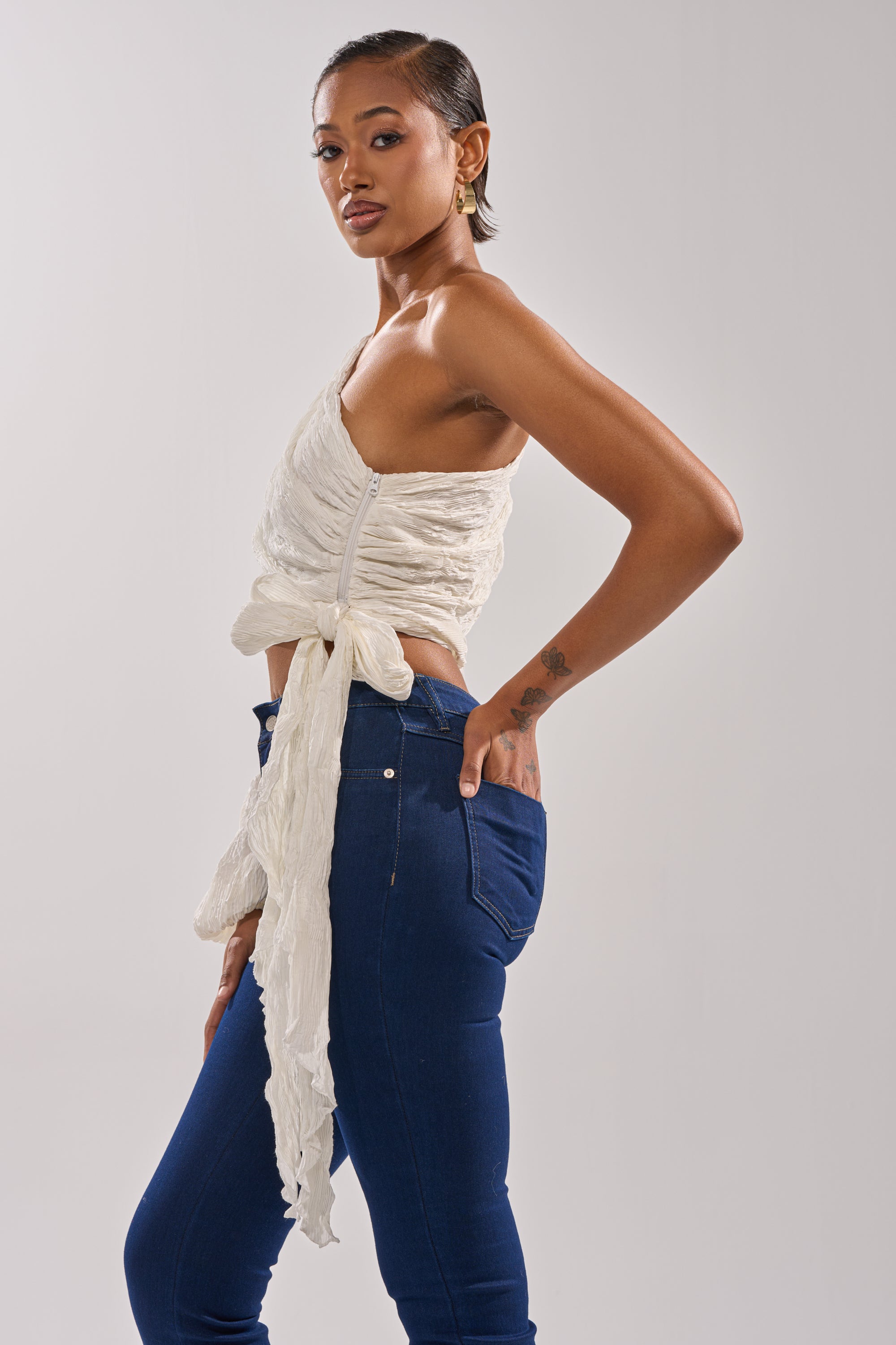 A woman with short hair and gold hoop earrings poses sideways against a plain background, wearing the GROOVY BABY SIDE TIE BLOUSE in a cropped fit, paired with high-waisted blue jeans. 4