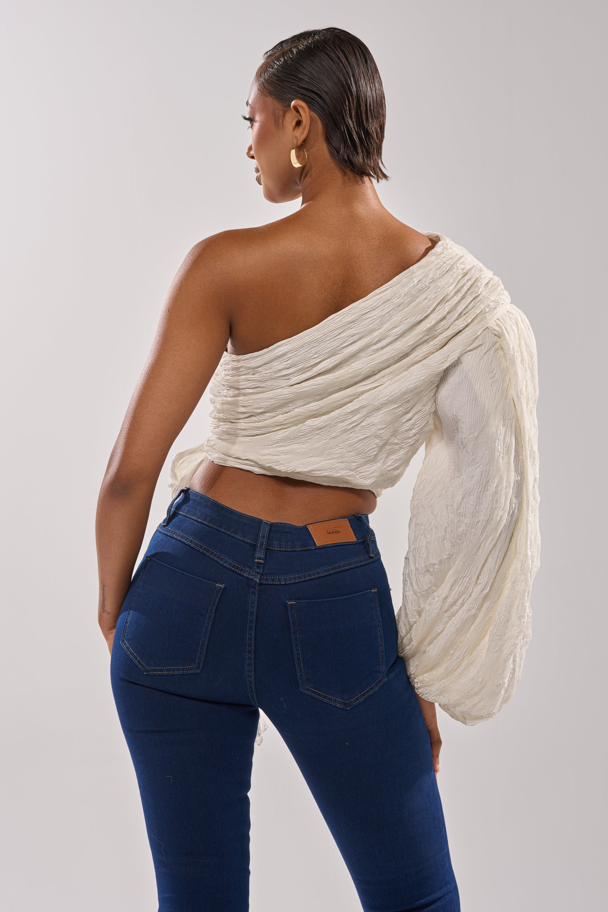 A woman with short hair and gold hoop earrings wears the GROOVY BABY SIDE TIE BLOUSE, a textured, off-the-shoulder white cropped top with one long billowy sleeve, paired with dark blue jeans against a plain light gray background. 5