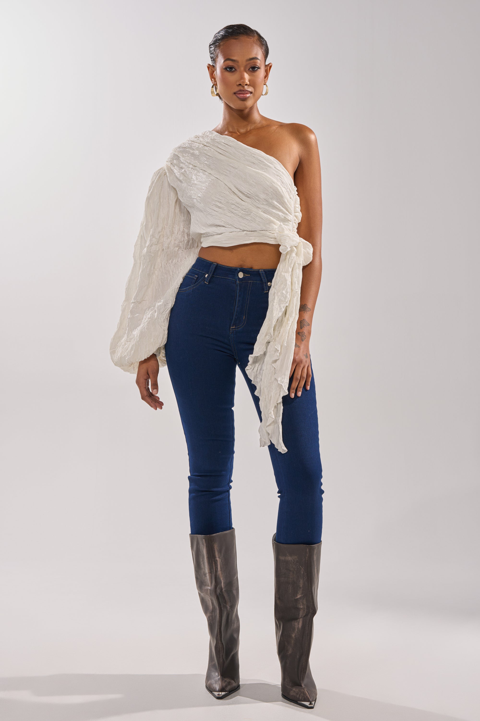 A woman stands confidently against a plain background, wearing the GROOVY BABY SIDE TIE BLOUSE—a white, one-shoulder cropped top—paired with dark blue skinny jeans and knee-high gray boots. 6