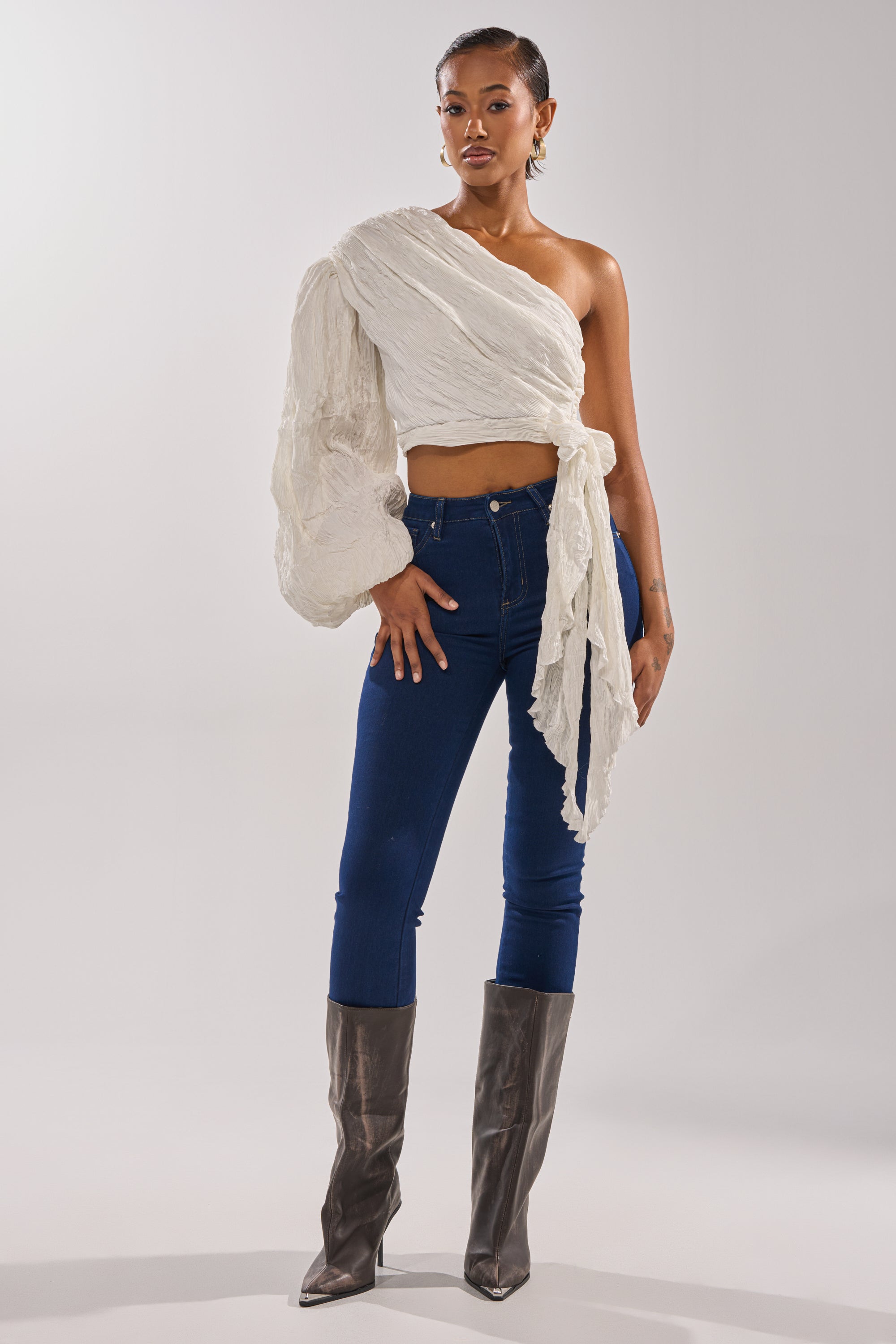 A woman stands confidently in the GROOVY BABY SIDE TIE BLOUSE, a textured white one-shoulder top with a voluminous sleeve, paired with blue skinny jeans and tall metallic gray boots against a plain light background. 7