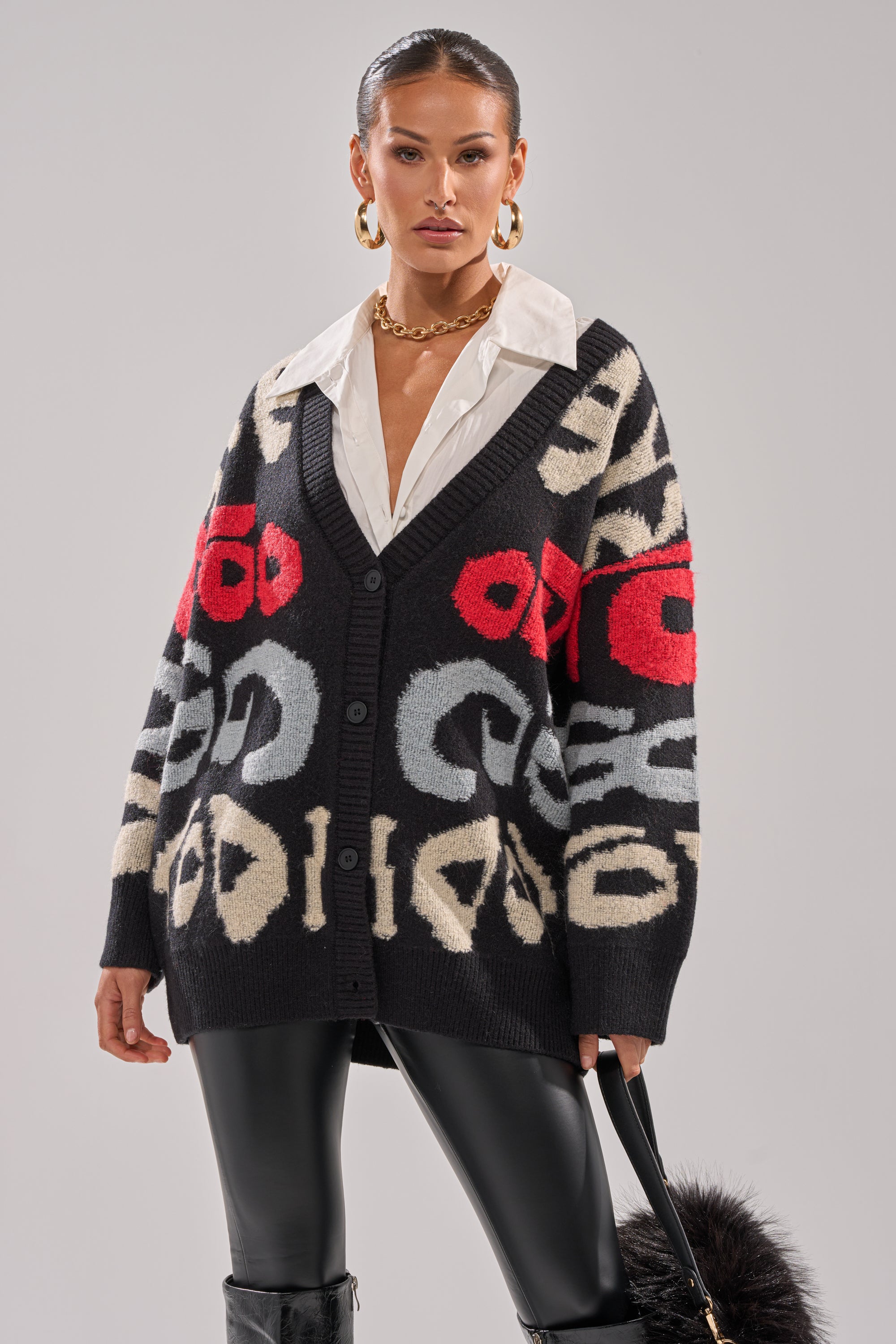 A woman styles the cozy GUESS WHO CARDIGAN, featuring bold red, white, and gray abstract patterns, over a white collared shirt and black leather pants. She finishes the outfit with a black faux fur accessory and gold hoop earrings.