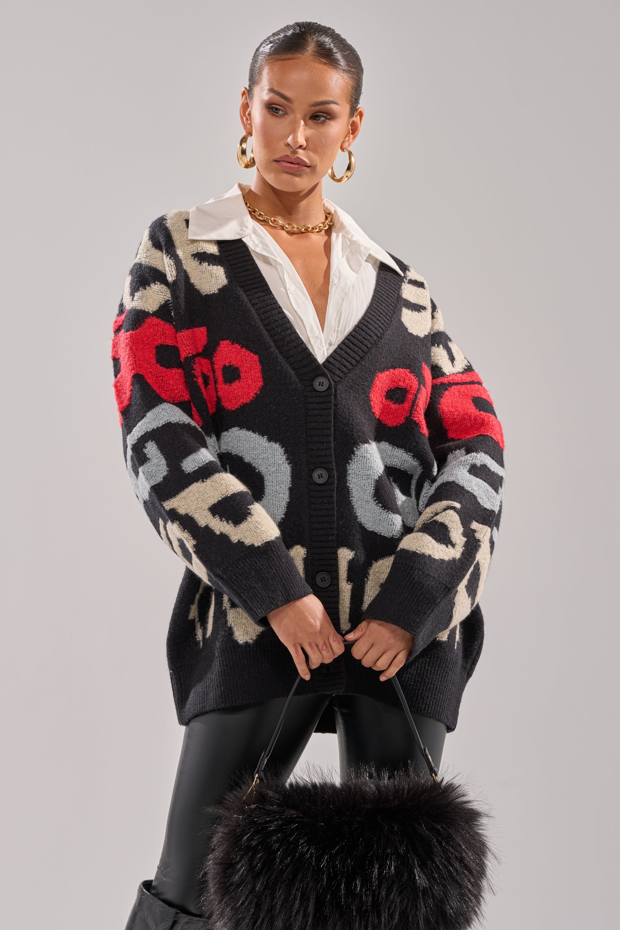 Woman in a cozy GUESS WHO CARDIGAN featuring bold red, white, and gray abstract patterns, styled with a white collared shirt, black pants, gold hoop earrings, chunky gold necklace, and a black faux fur handbag.