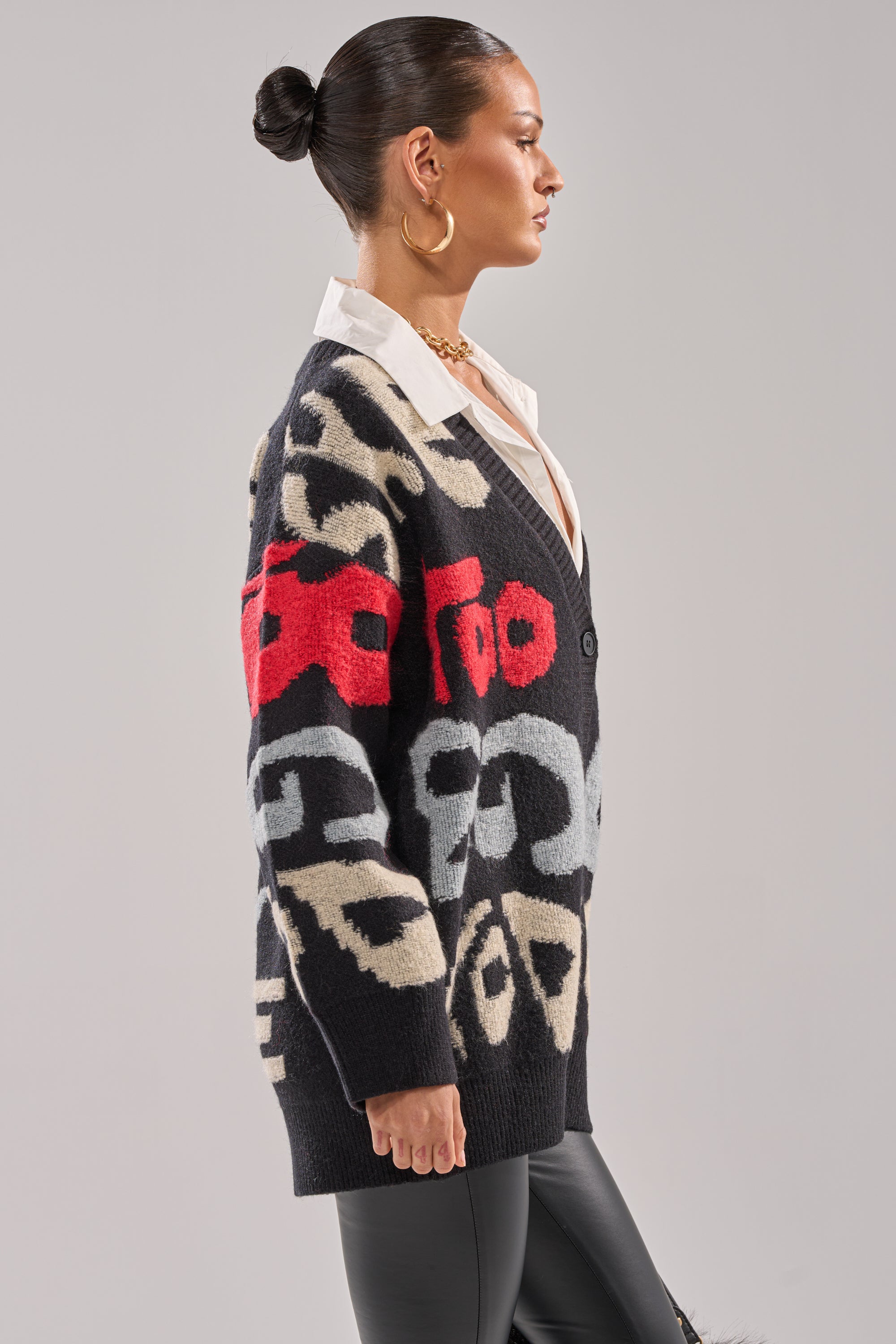 A woman in profile wears the GUESS WHO CARDIGAN—a black knit cardigan with red, cream, and gray abstract patterns—over a white collared shirt, gold hoop earrings, and black leather pants against a plain background.