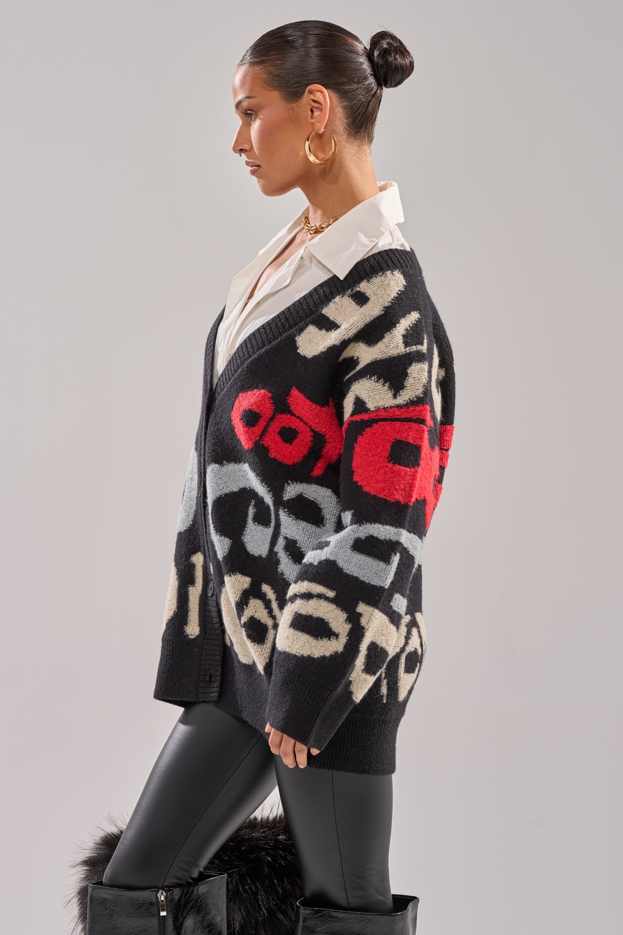 A woman with her hair in a bun wears a white collared shirt, the GUESS WHO CARDIGAN featuring abstract red, beige, and gray patterns, black leather pants, and black fur-trimmed boots while standing in profile against a light background.