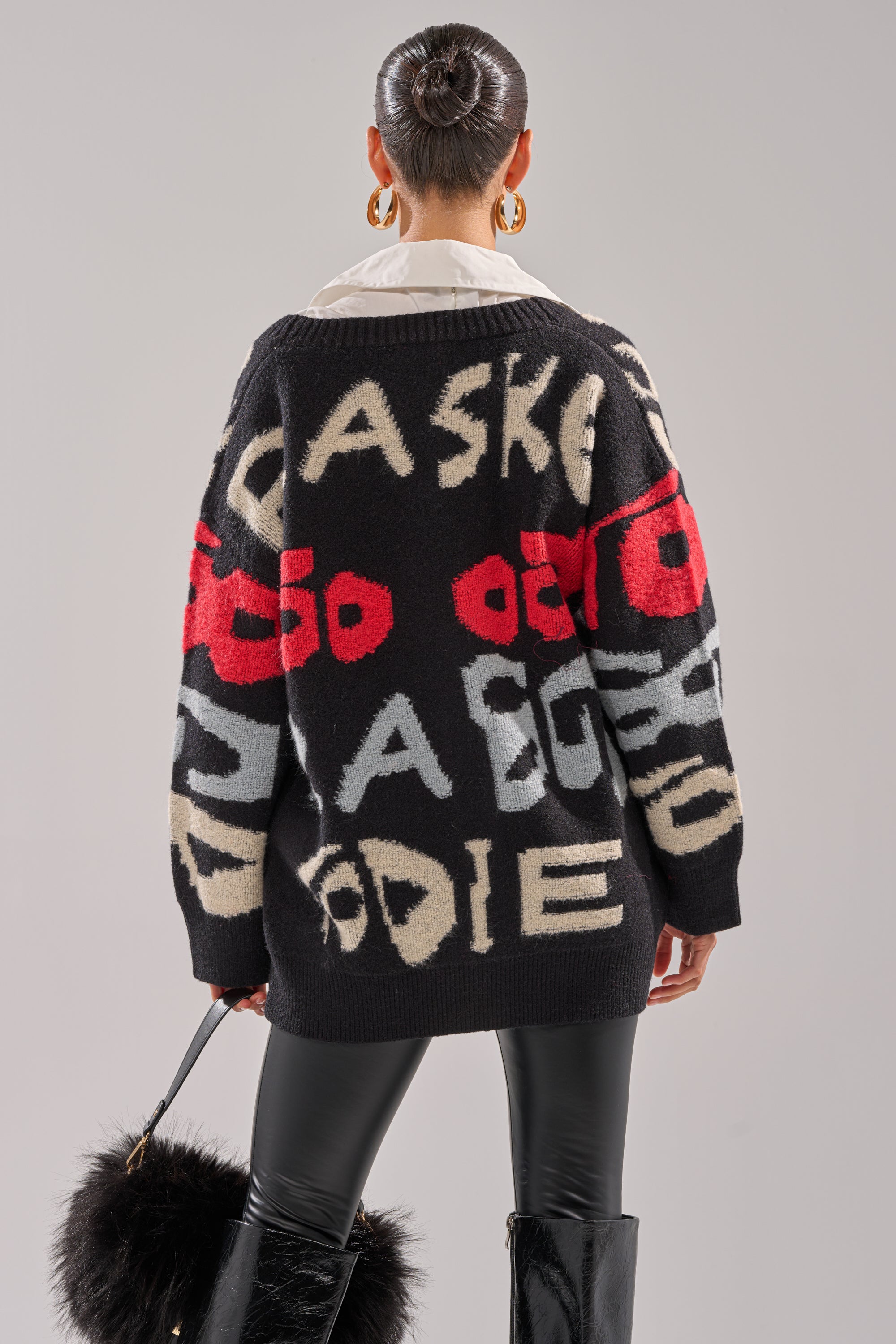 A woman with a neat bun, facing away, wears the GUESS WHO CARDIGAN in black with bold beige, red, and gray abstract text graphics, paired with black leggings, knee-high boots, and holds a black fur bag.