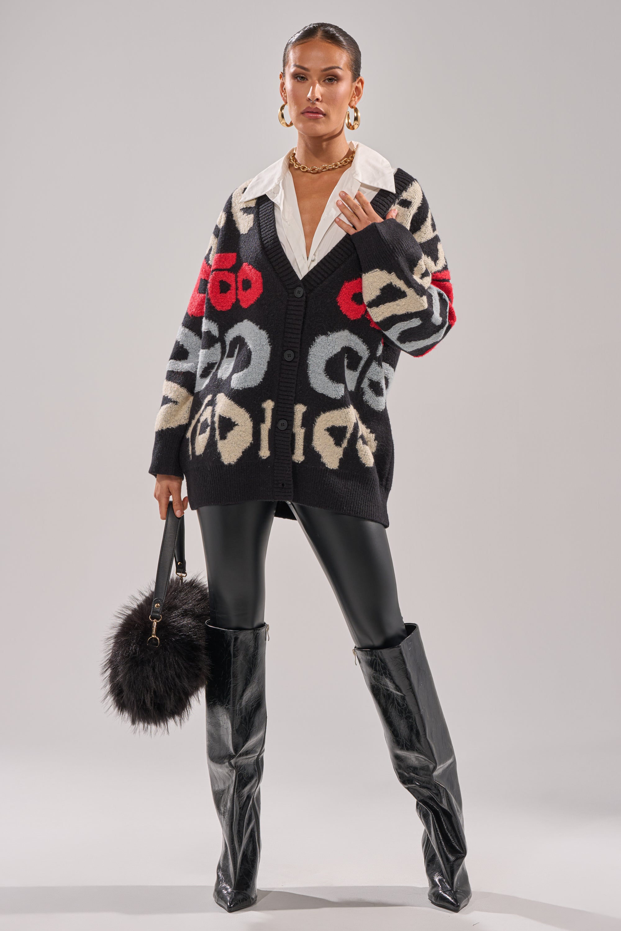 A woman confidently wears the GUESS WHO CARDIGAN—an abstract, multicolor knit—over a white collared shirt with black leather leggings, knee-high boots, gold hoops, and a furry black bag against a plain backdrop.