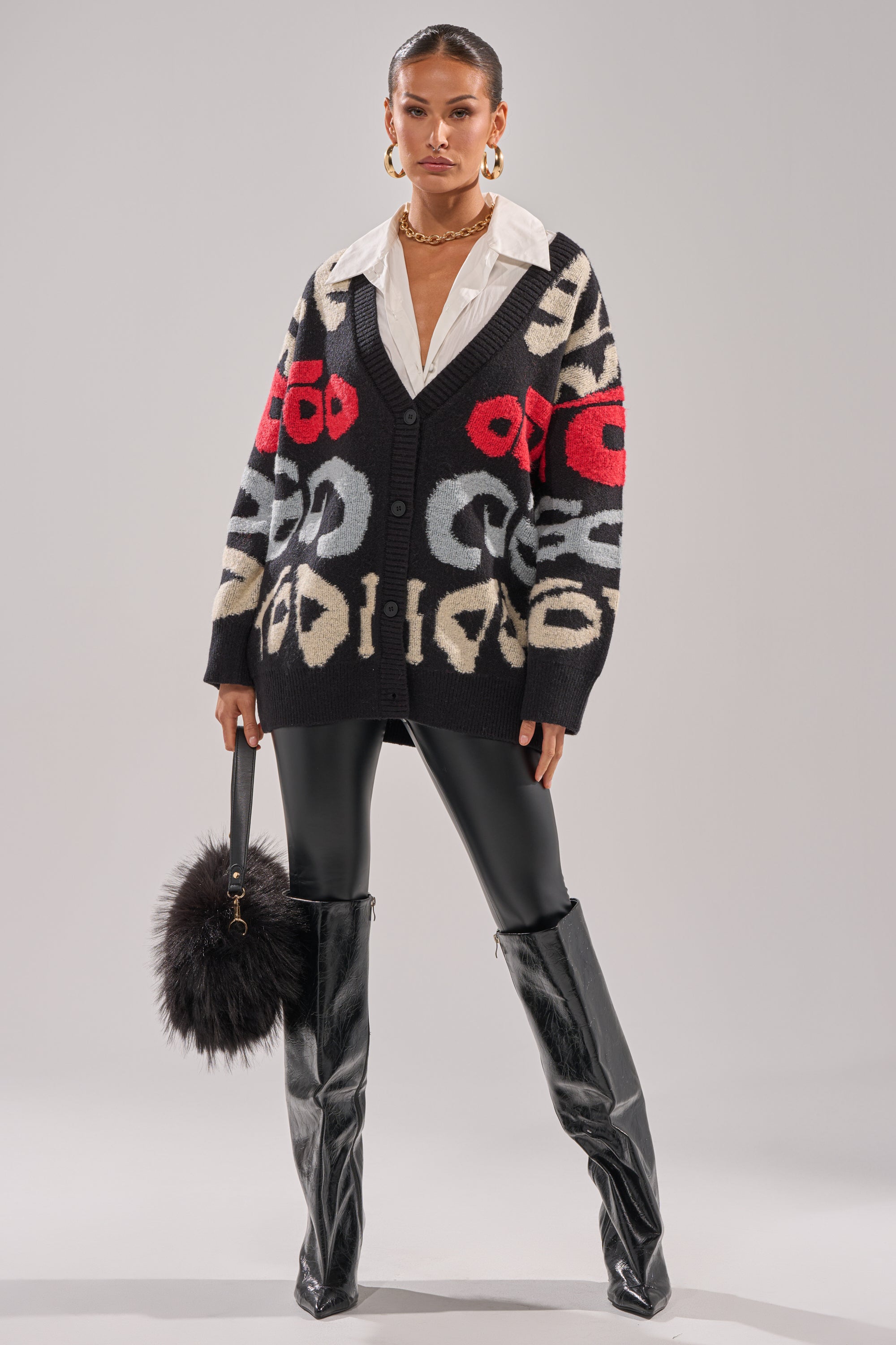 A woman models the GUESS WHO CARDIGAN with a white collared shirt, black leggings, knee-high black boots, hoop earrings, and a black furry handbag against a plain gray background.