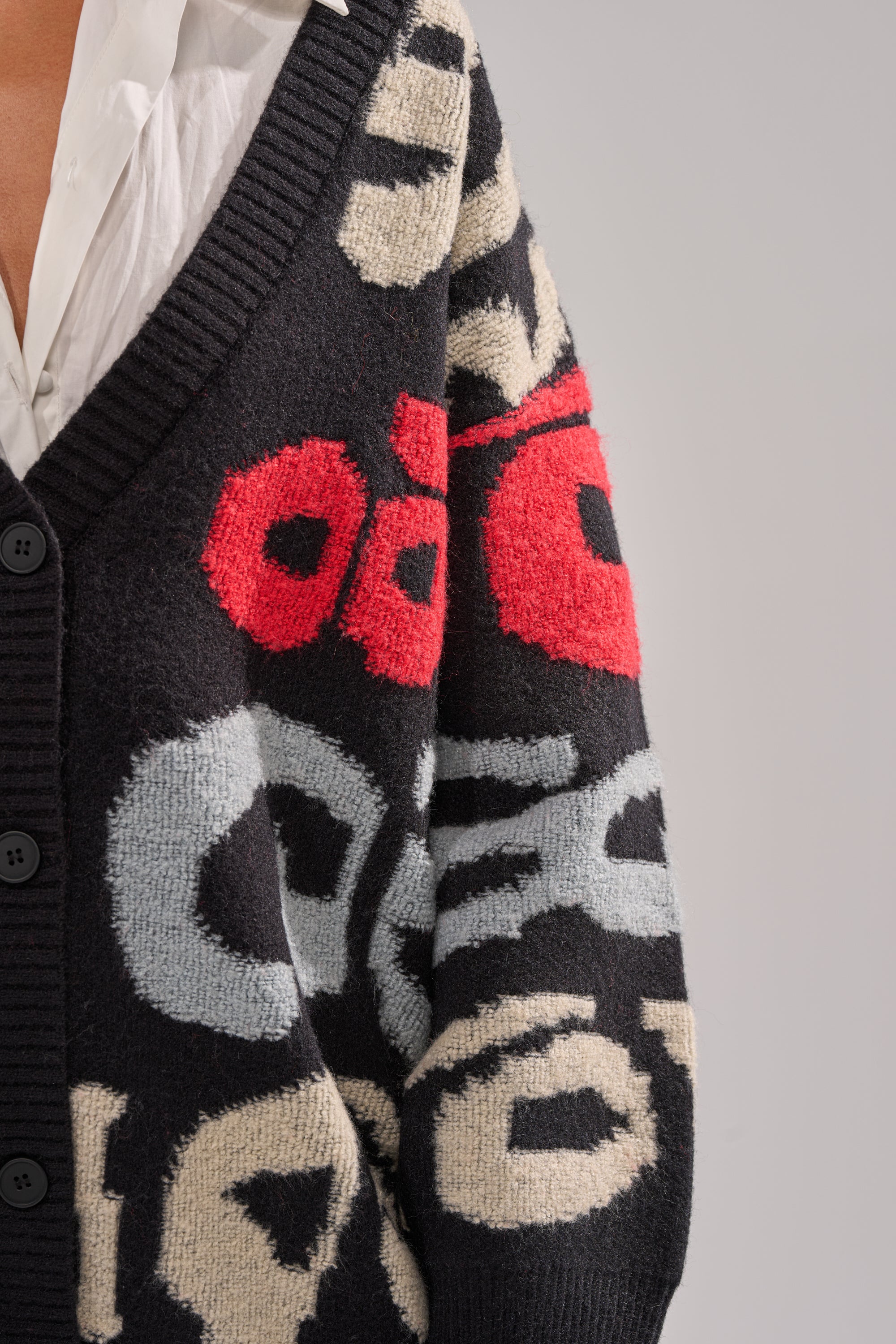 Close-up of a person wearing the GUESS WHO CARDIGAN, featuring bold abstract knit patterns in red, white, and gray over a white collared shirt. The photo shows the chest and arm against a simple background.