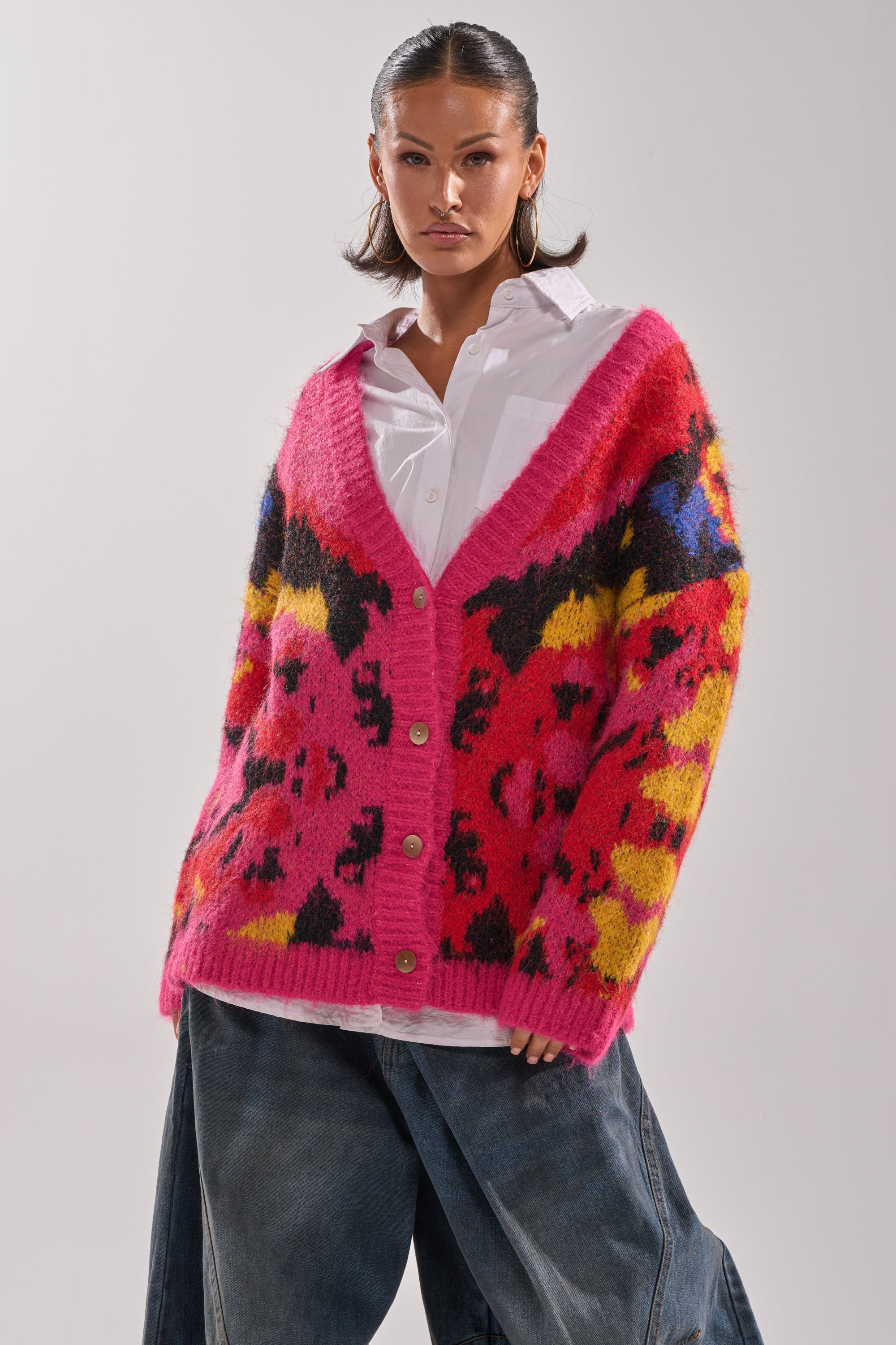 A woman pairs a loose white shirt and wide-leg denim jeans with the COZY VIBES CARDIGAN, featuring vibrant pink, black, yellow, and blue abstract patterns. She stands confidently against a plain, light background.