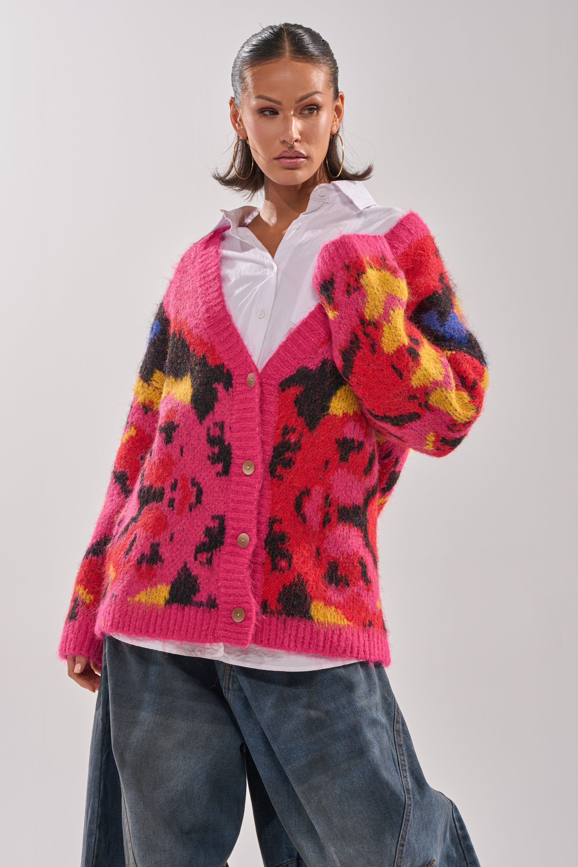 A woman poses confidently in the COZY VIBES CARDIGAN—a bright pink, fuzzy knit with an abstract multicolored pattern—styled over a white button-up and loose blue jeans against a plain background.