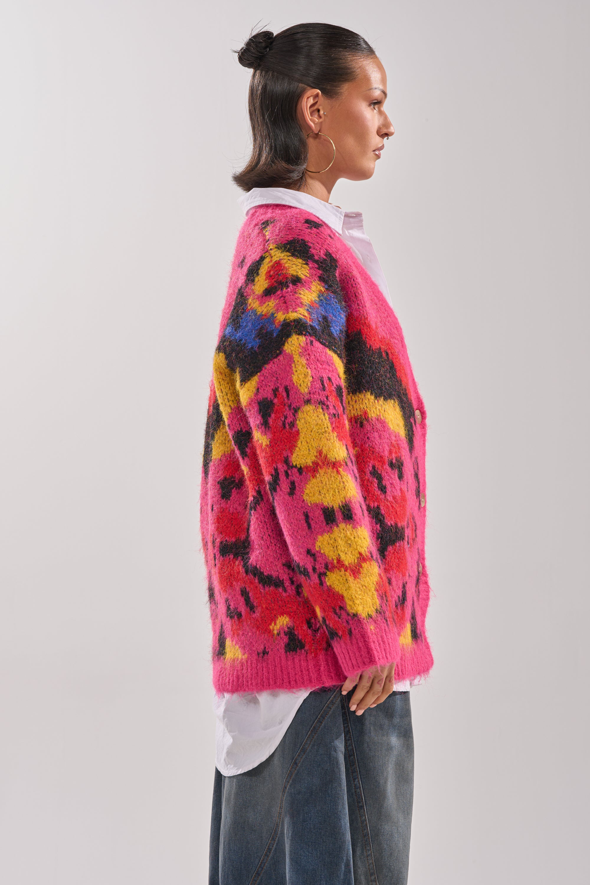 A woman in profile wears the COZY VIBES CARDIGAN, a fuzzy knit with abstract yellow, blue, and black designs, layered over a white shirt and wide-leg blue jeans against a plain light background.