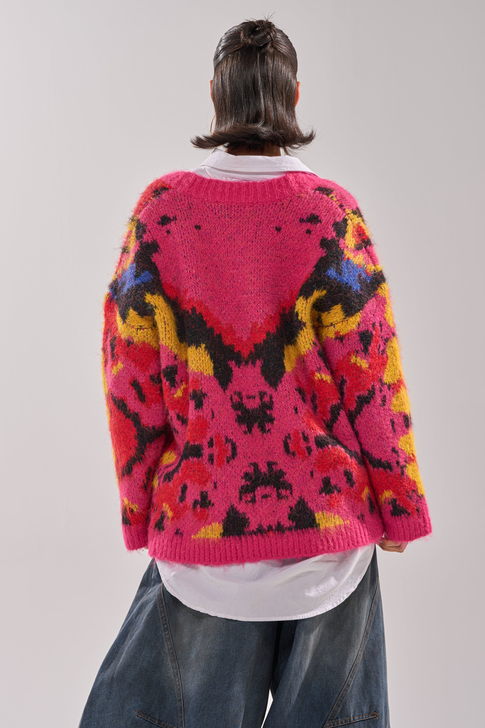 A person with shoulder-length hair stands with their back to the camera, wearing the COZY VIBES CARDIGAN in bright pink with yellow, black, and red abstract patterns over a white shirt and wide-leg blue jeans.