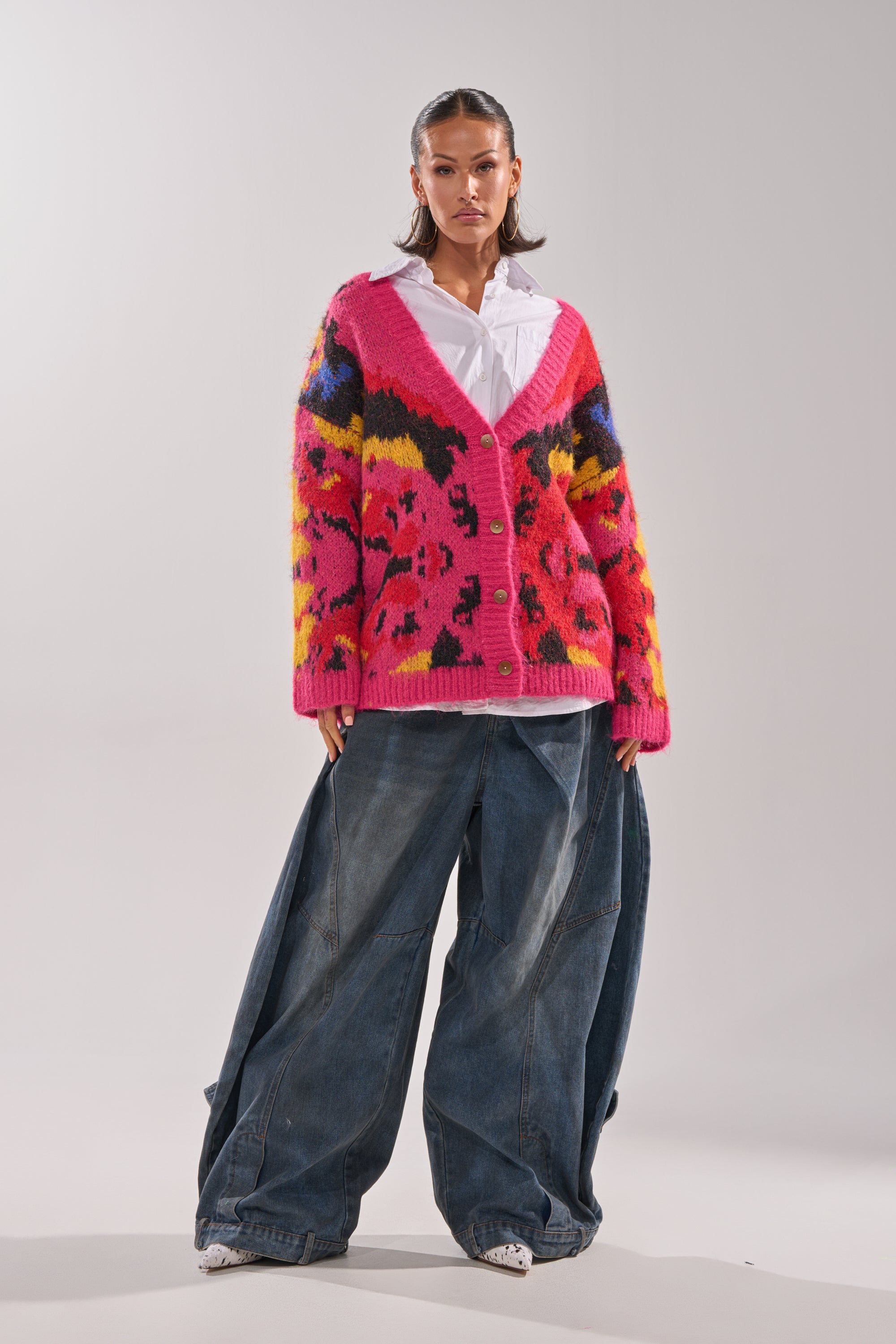 A woman stands confidently in a white shirt and baggy blue jeans, layered with the COZY VIBES CARDIGAN—an oversized knit featuring bold, colorful patterns. The plain background showcases her stylish, eclectic look.
