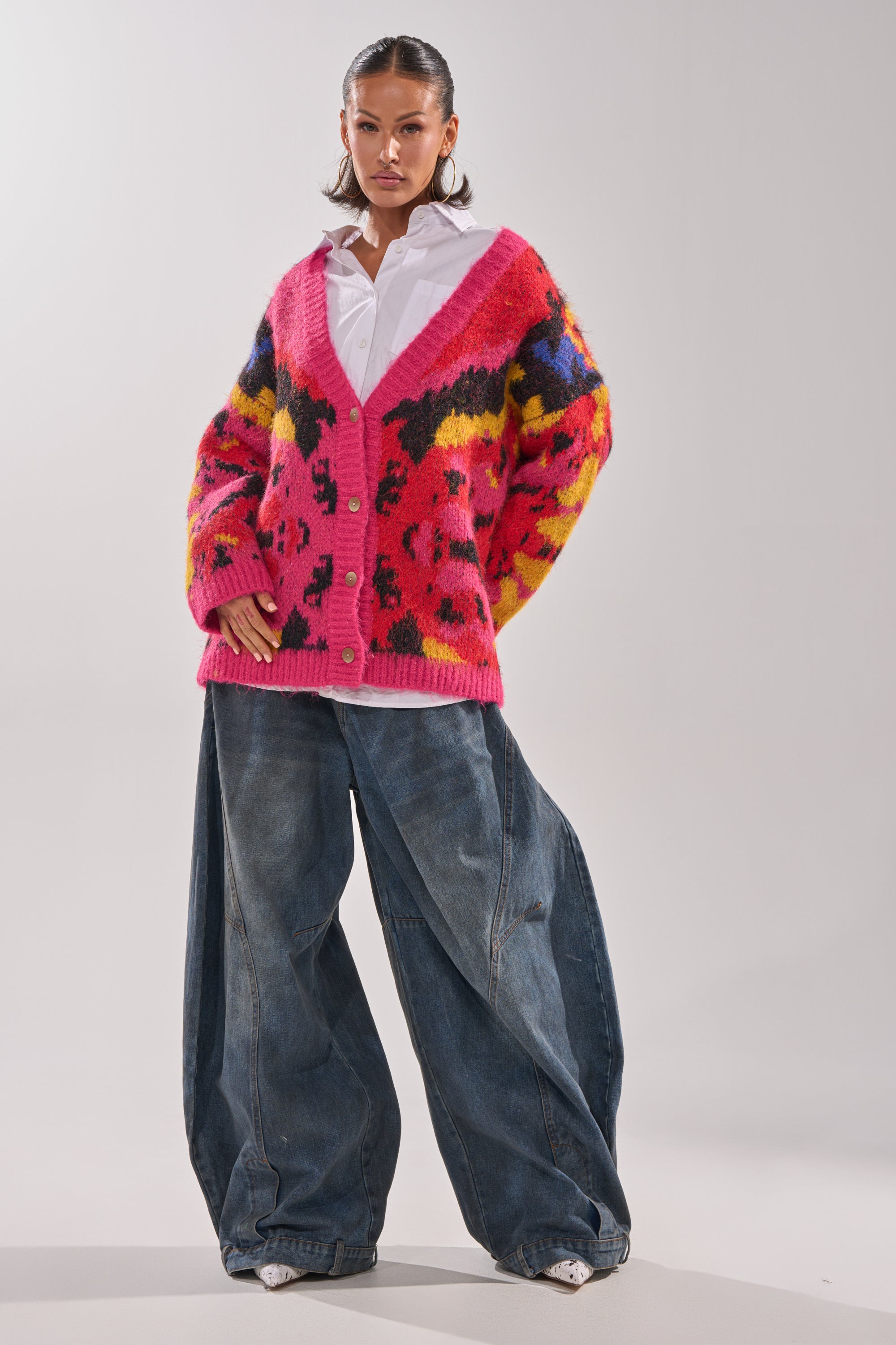 A woman confidently wears the COZY VIBES CARDIGAN, an oversized, multi-color jacquard piece with a bold pattern, styled over a white collared shirt and wide-leg blue jeans against a plain light background.