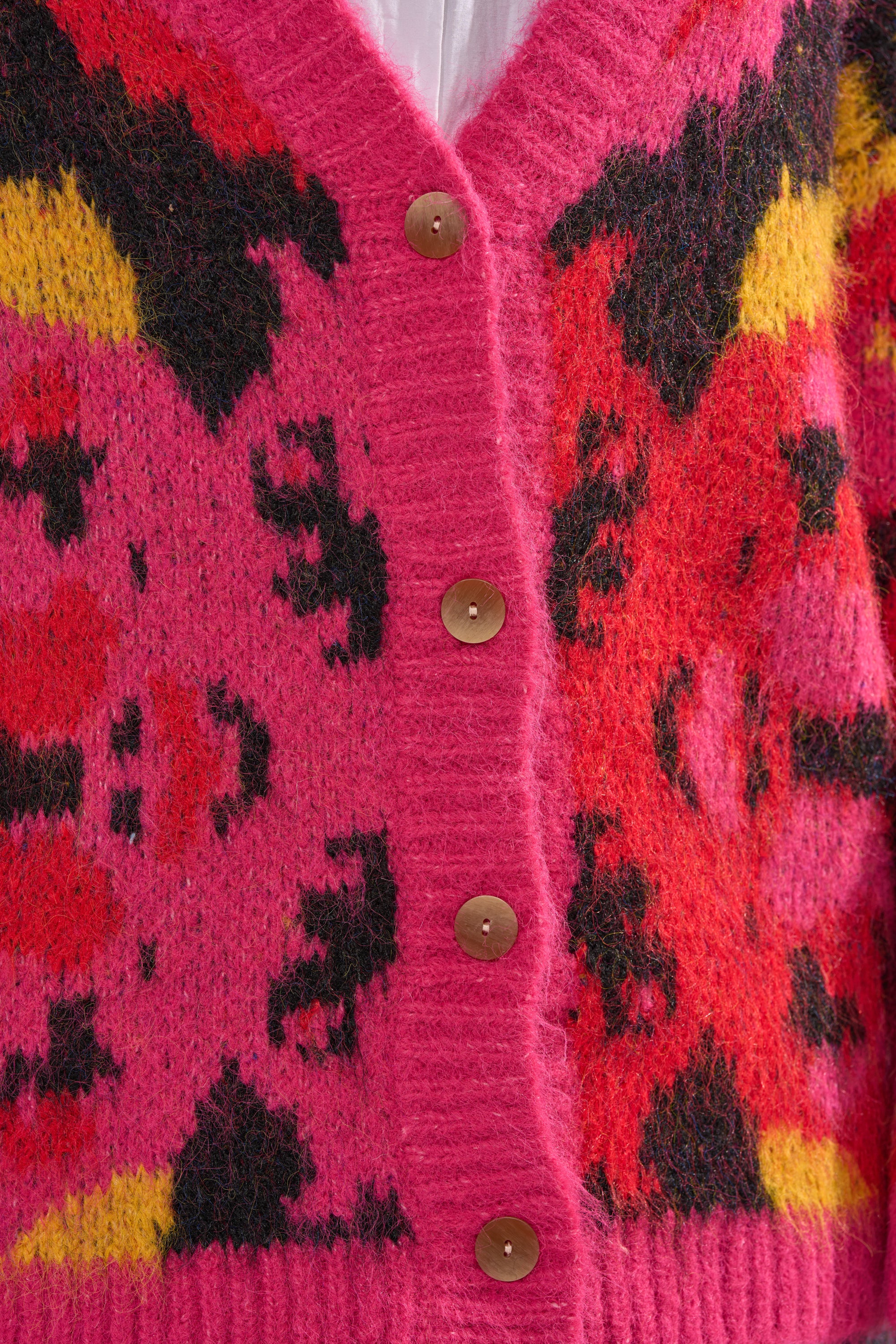 Close-up of the COZY VIBES CARDIGAN, a fuzzy pink knit with red, black, and yellow abstract patterns. Features four brown front buttons and ribbed trim—ideal for adding cozy charm to any outfit.
