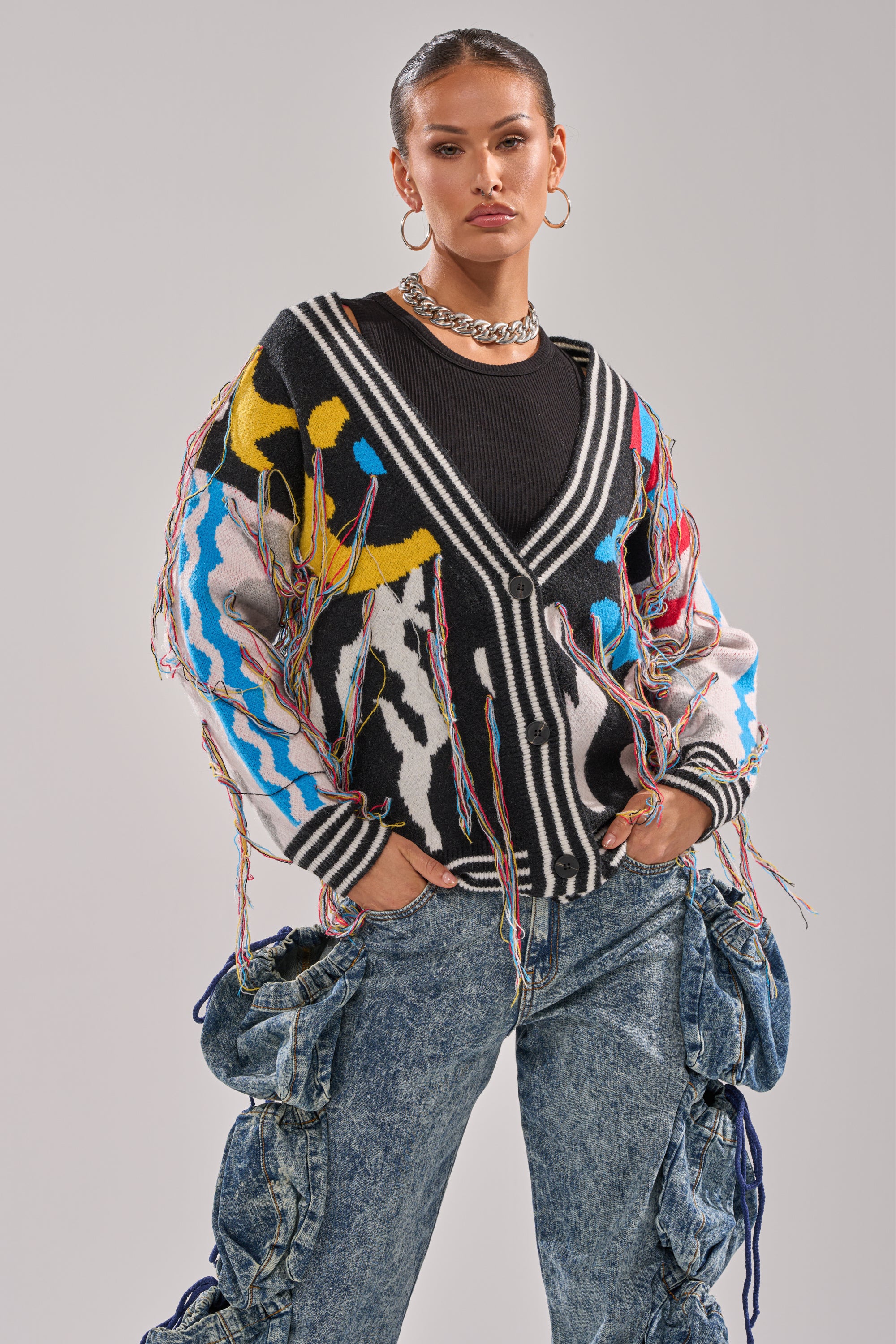A woman poses confidently in the SWEATER WEATHER CARDIGAN, a colorful oversized fringe cardigan, paired with a black top, chunky necklace, hoop earrings, and baggy blue jeans with ruffled sides against a light gray background.