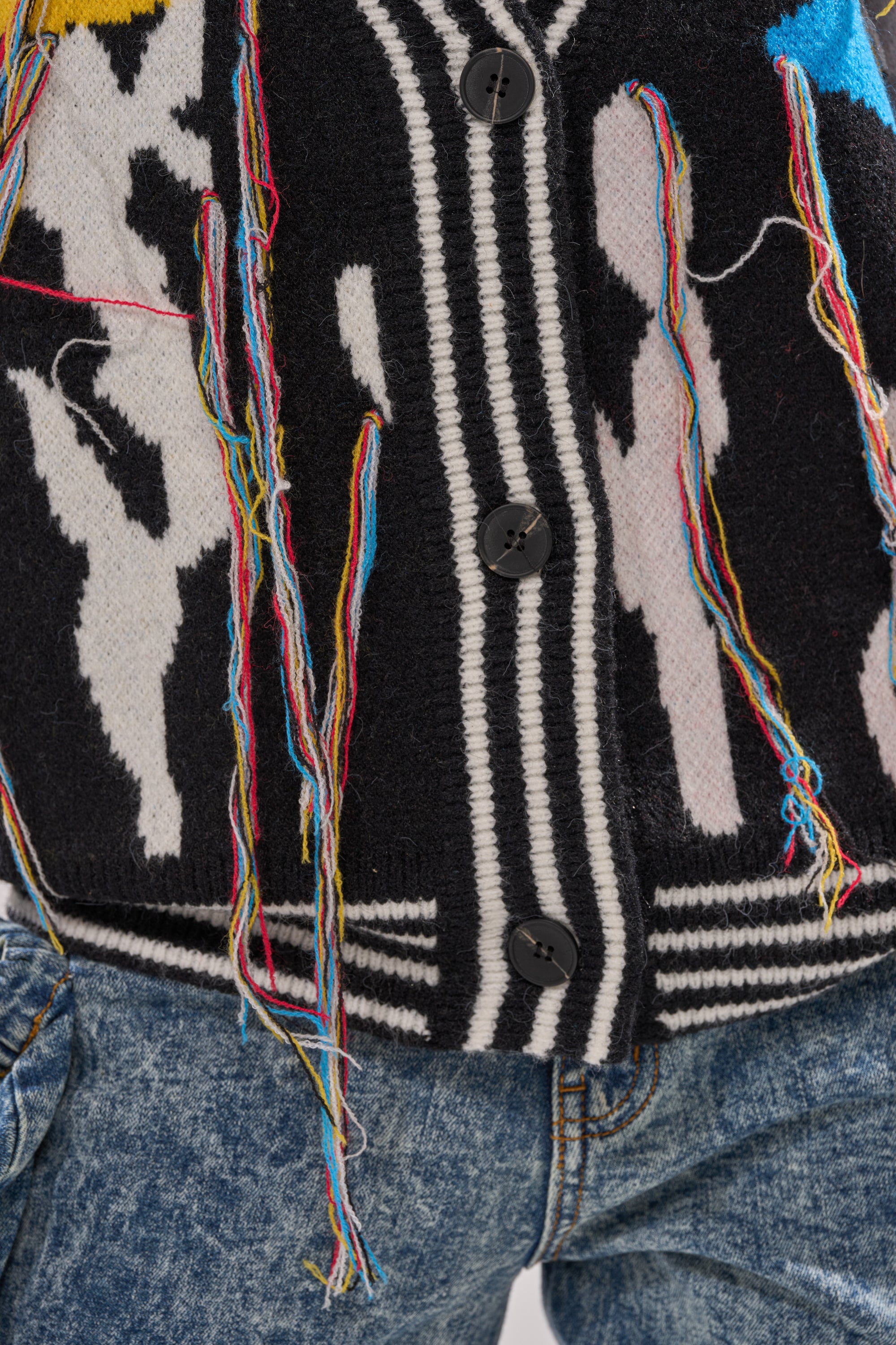 The SWEATER WEATHER CARDIGAN is a cozy black and white knit with colorful loose threads and black buttons, styled with blue denim jeans—ideal for sweater weather.