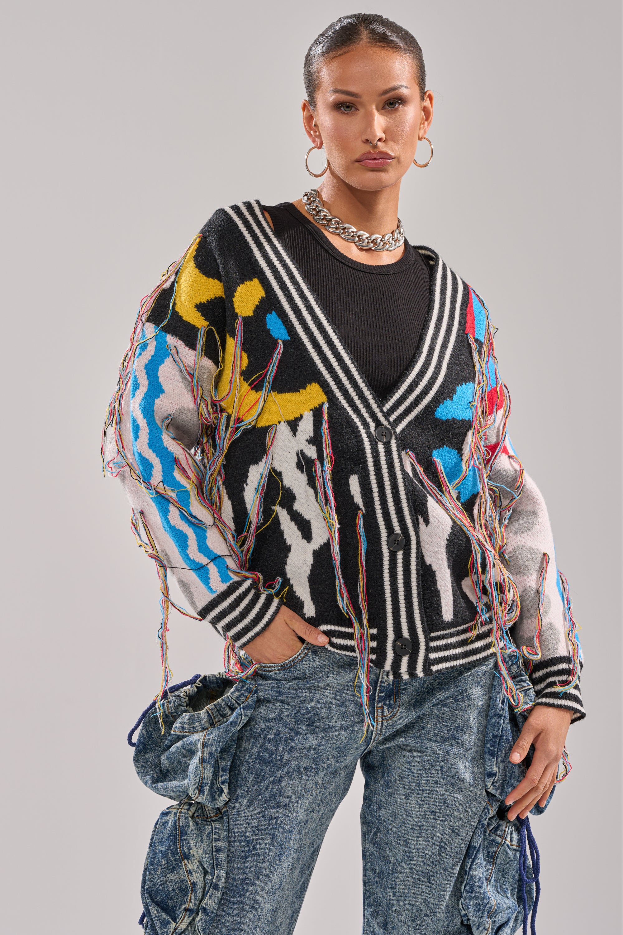 A woman poses confidently with her hair pulled back, wearing the colorful SWEATER WEATHER CARDIGAN with fringe details over a black top, styled with a chunky chain necklace and blue jeans featuring large pockets.