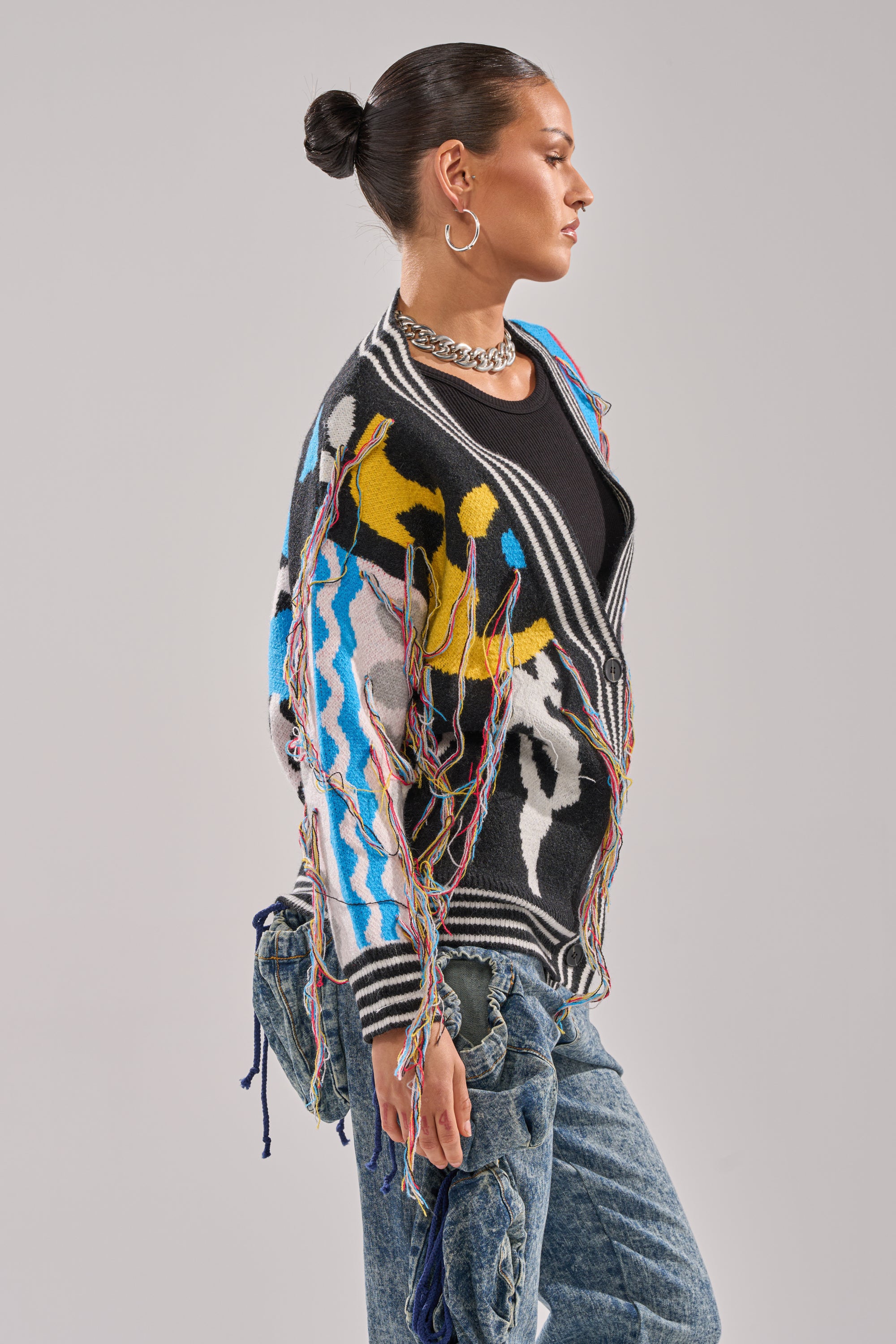 A person with dark hair in a bun wears the SWEATER WEATHER CARDIGAN—featuring colorful patterns and fringe—over a black top with blue jeans, hoop earrings, and a chunky chain necklace, standing in profile against a plain background.