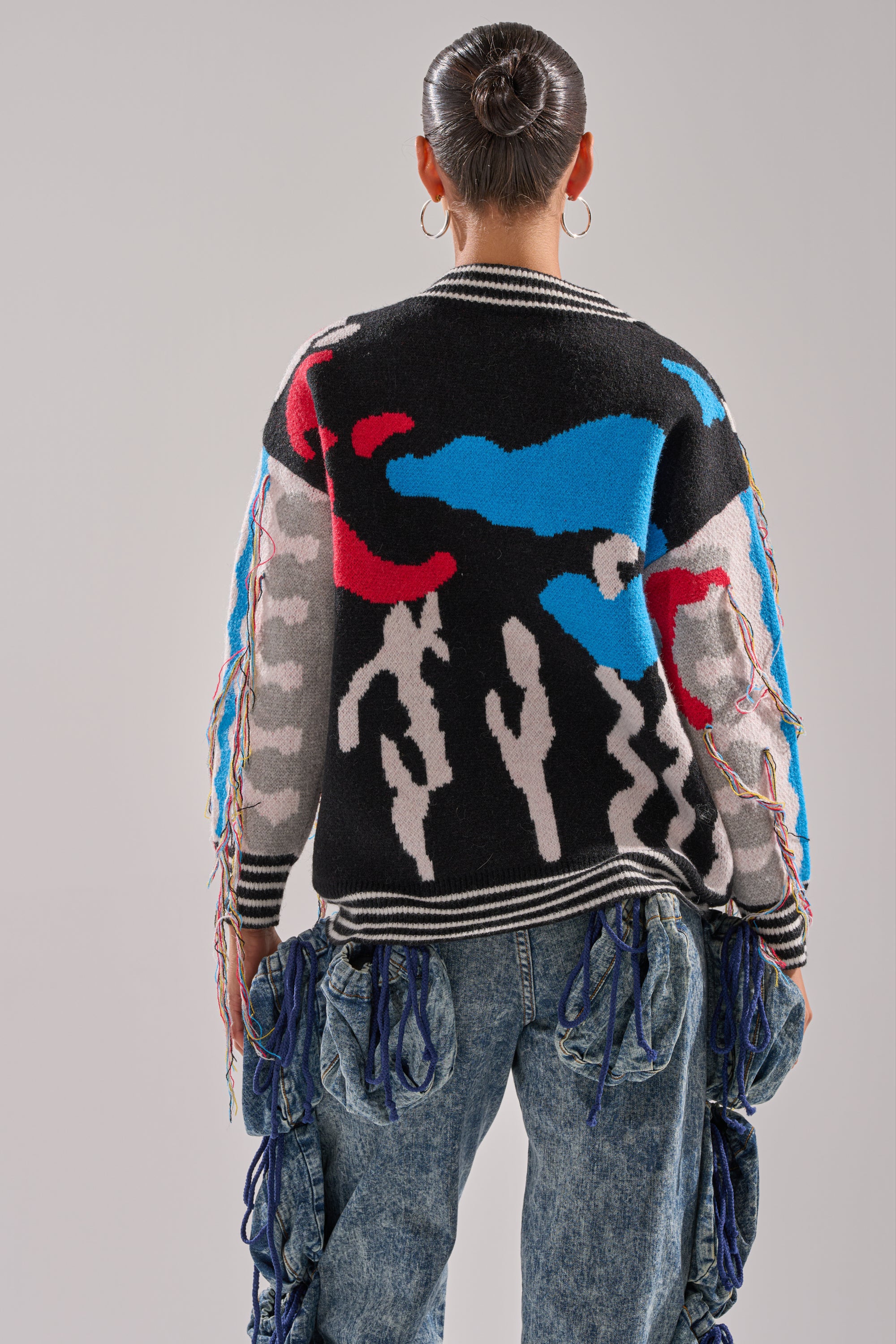 A person wearing the SWEATER WEATHER CARDIGAN, styled with a bun, large hoop earrings, and blue jeans with tassels, stands with their back to the camera against a plain background.