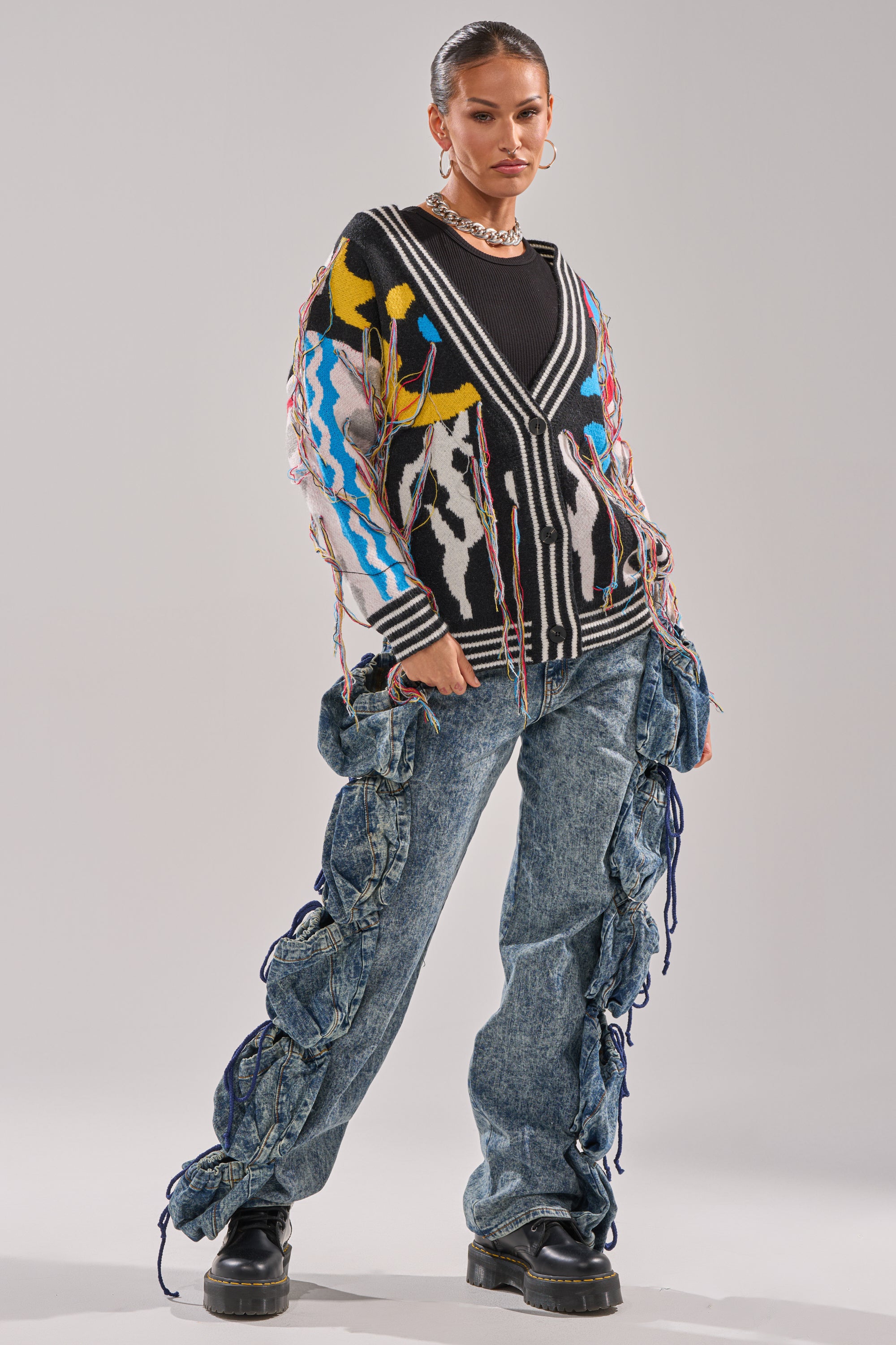 A woman stands confidently in the SWEATER WEATHER CARDIGAN, styled with loose light-wash jeans featuring large ruched sections tied with blue strings. Chunky black shoes and hoop earrings complete her look.