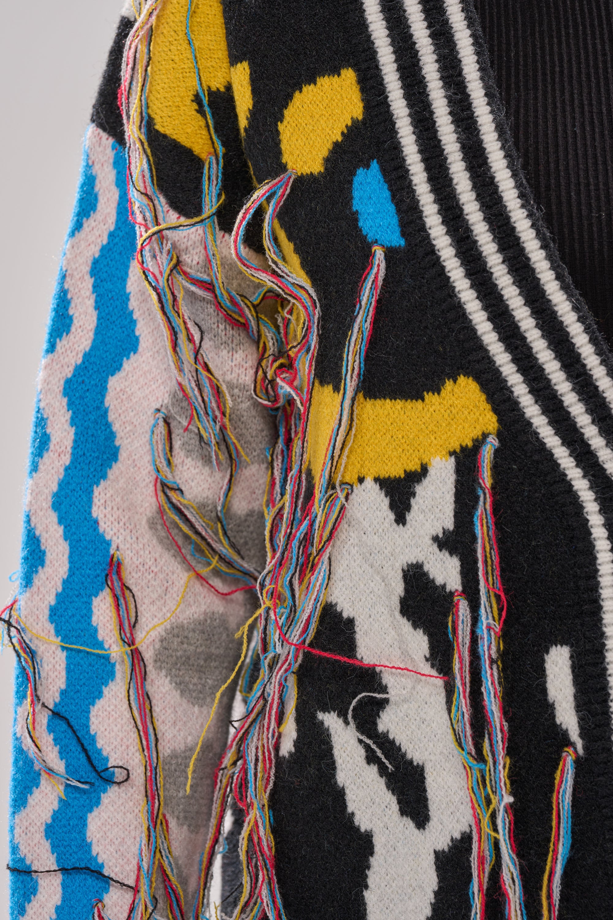 A close-up of the SWEATER WEATHER CARDIGAN’s sleeve displays loose, multicolored threads and abstract patterns in black, yellow, blue, gray, and white, finished with a black and white striped trim near the collar.