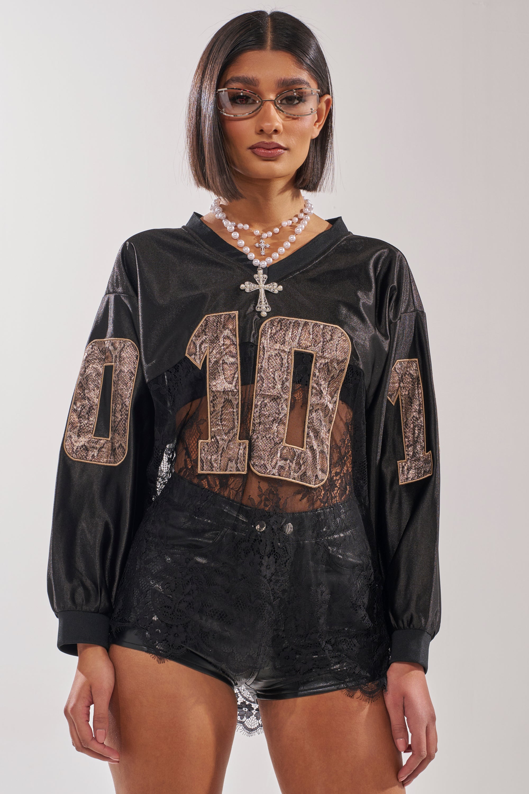 A woman wearing sunglasses, a pearl necklace with a cross pendant, the ROCKY LACE JERSEY featuring "0101" in snakeskin print, and black lace shorts stands against a plain light background for a streetwear-ready look. 0