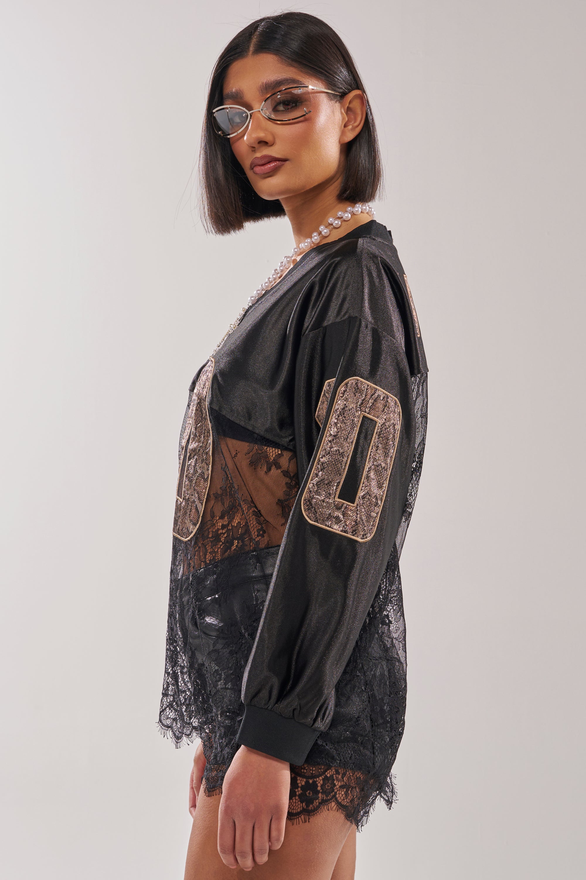 A woman with straight dark hair, clear glasses, a pearl necklace, and an oversized ROCKY LACE JERSEY with "00" on the sleeves poses against a plain light background for a streetwear-inspired look. 4
